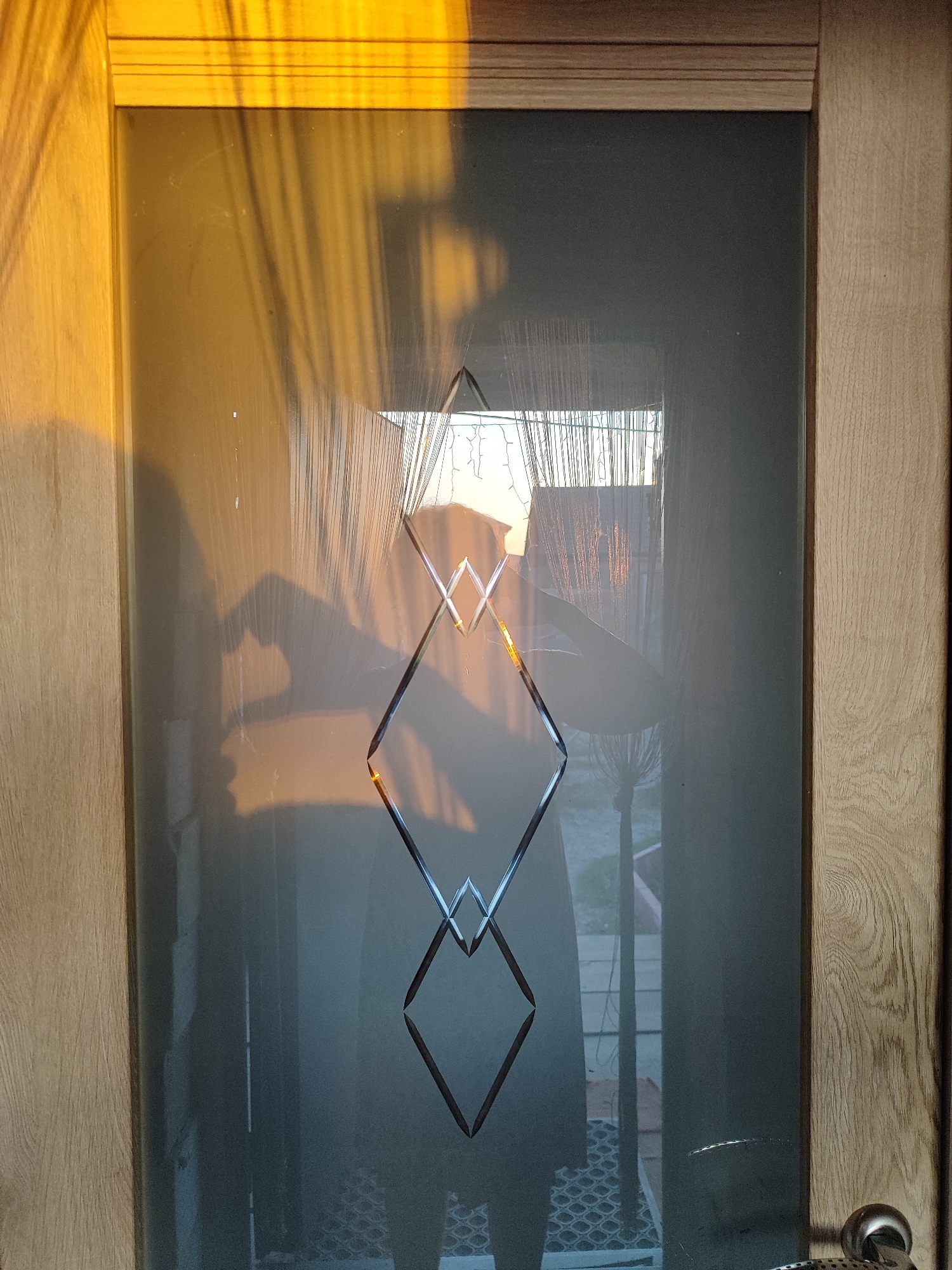 This image shows a close-up of a wooden door with a glass pane featuring a decorative etched diamond pattern. The glass is partially obscured by shadows and reflections, suggesting it is daytime and light is streaming in. A person's shadow is visible on the left side of the glass, with their hand appearing to be in a heart shape. The background seen through the glass is blurry and appears to be an outdoor setting, possibly a yard or street with some fencing and vegetation. The lower right corner of the image shows a portion of a door handle. The photograph was taken in Marusino, Russia.