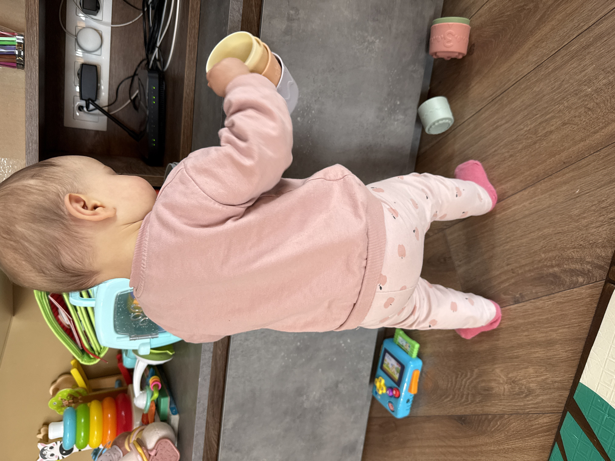 A young child wearing a pink top and patterned pants is standing on a wooden floor next to a gray surface. The child is holding a stack of cups in their hand. Several toys are on the floor, including a blue toy with buttons, a toy stack of rings, and a set of cups. Additional toys are located in the lower-left corner of the frame. Electrical outlets and cords are visible near the child's head. The location is unknown.