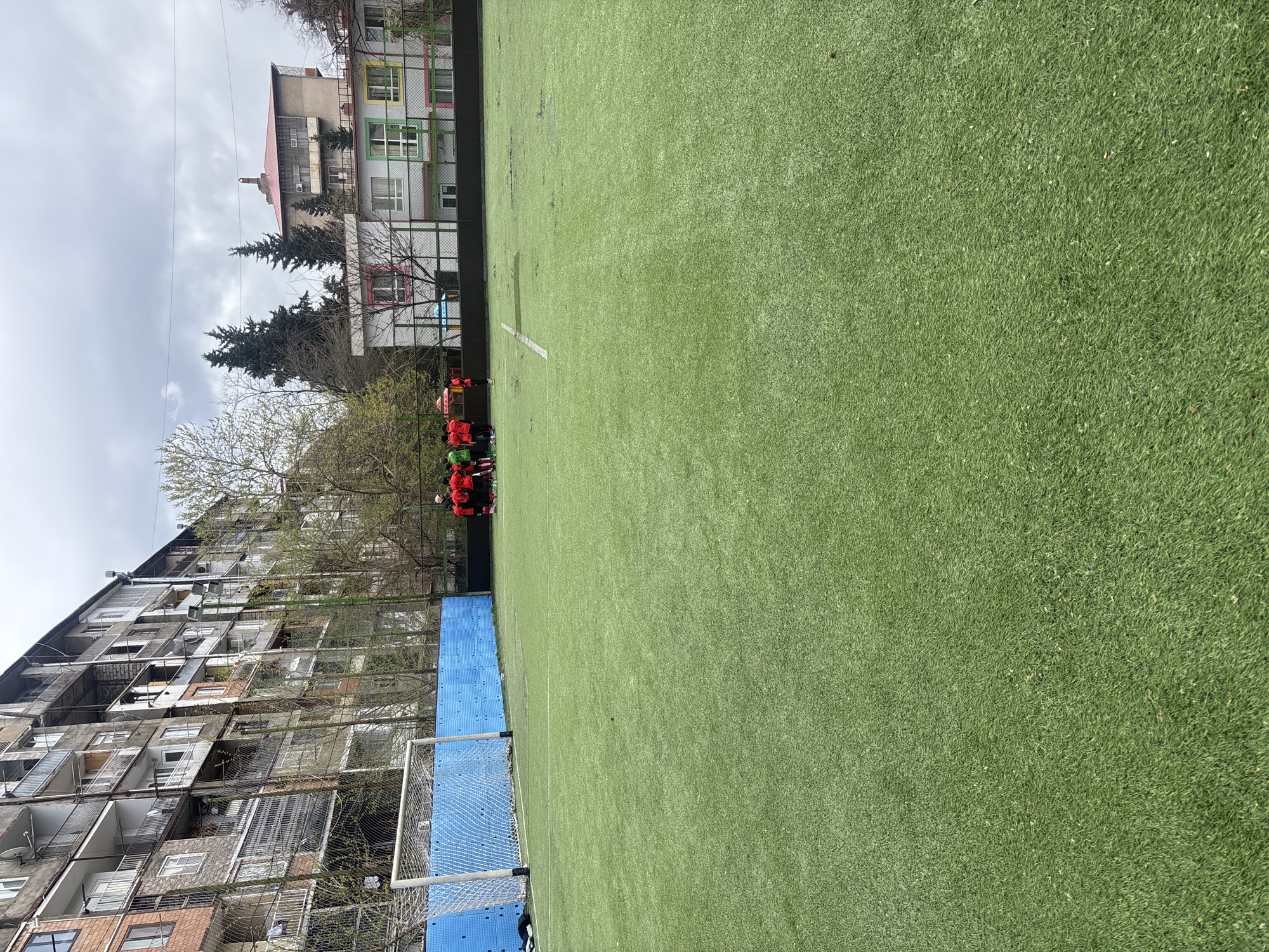 The image depicts a small artificial turf sports field in Tbilisi, Georgia. A group of people wearing red athletic apparel are huddled at one edge of the field. Adjacent to the field are buildings of varied architecture, including a multi-story residential structure with balconies and a building with brightly colored window frames. There is a blue-framed soccer goal with white netting visible along one sideline of the sports field. The sky is overcast.