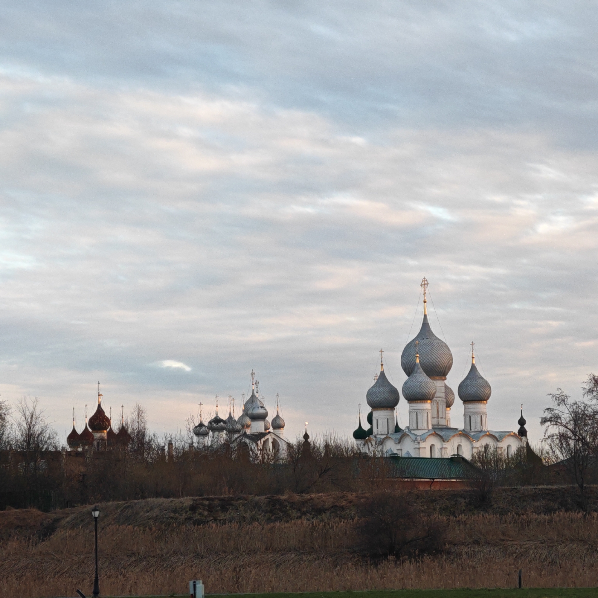 The image depicts a cityscape in Rostov, Russia, showcasing an Orthodox church complex. The foreground features dry vegetation and a low embankment. Behind this, a cluster of buildings with onion-shaped domes is visible. Some domes are silver, while others are red. The sky is overcast with a mix of white and gray clouds. A lamppost is visible on the left side of the image. The scene suggests a rural or suburban environment. There are no people visible in the image.