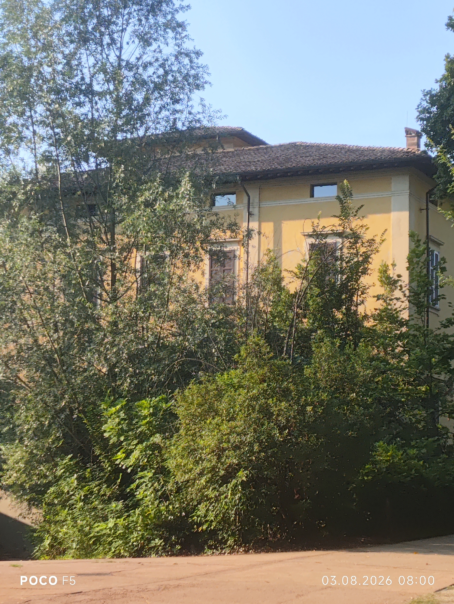 A sunlit, over-the-shoulder shot shows a traditional Italian villa partially obscured by lush, green trees and foliage. The villa, painted a warm yellow, features a red tiled roof and dark-shuttered windows. A lone, red-bricked chimney stands tall against the clear blue sky. The foreground consists of a dusty, reddish-brown path leading up to the property, suggesting a rural or semi-rural setting outside the town of Collecchio, Italy. The image is dated 03.08.2026 at 08:00 and displays "POCO F5" branding, indicating it was taken with a Poco F5 smartphone. There are no people or animals present in the scene.