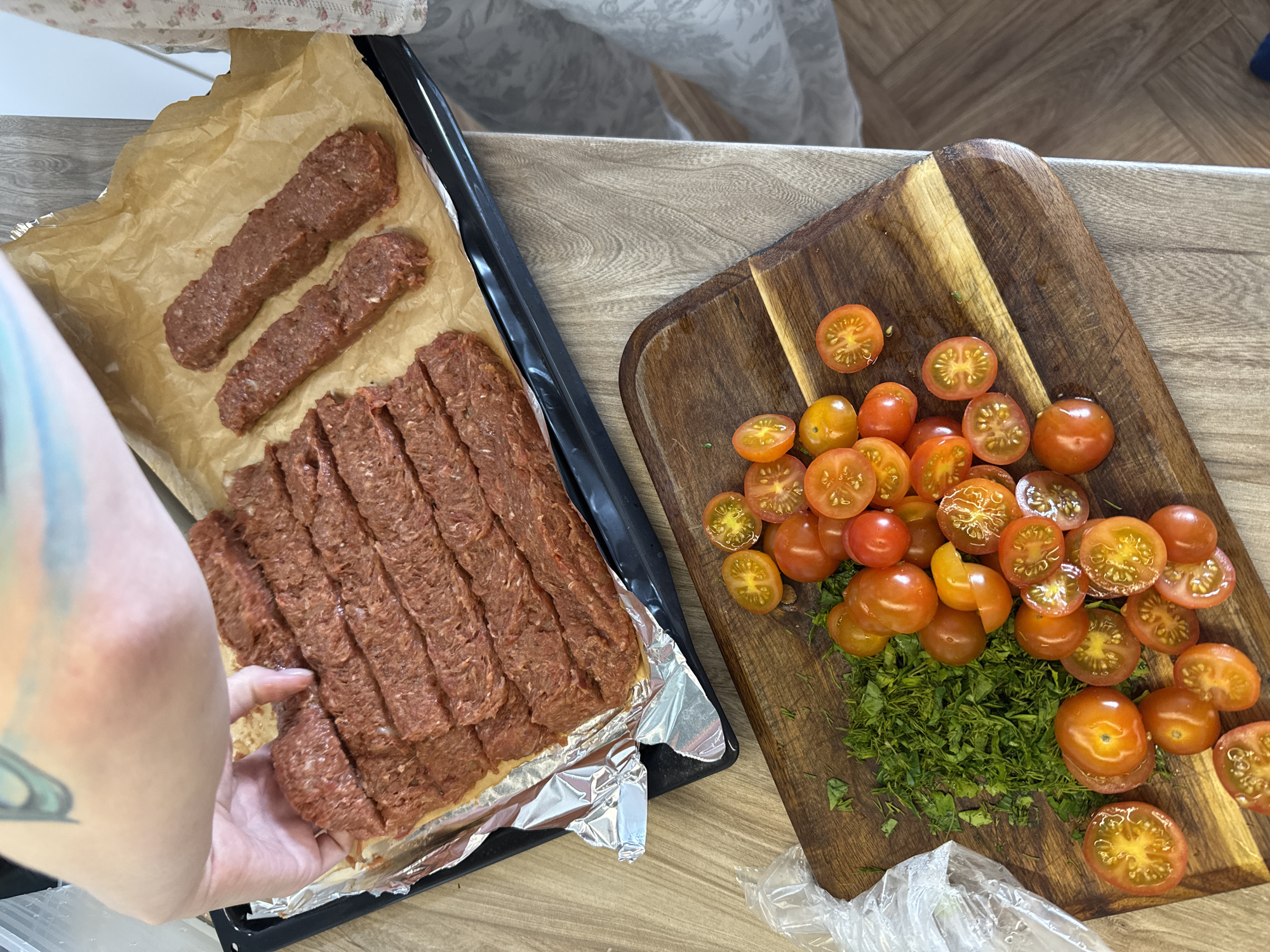 A person is preparing a meal, consisting of raw ground meat formed into elongated rectangular shapes and sliced into segments, alongside halved cherry tomatoes and chopped greens on a wooden cutting board. The meat is arranged on a baking sheet lined with parchment paper and aluminum foil. The scene appears to be indoors on a kitchen countertop with a light-colored wood grain pattern. It is not possible to confirm the location as Krasnoyarsk, Russia based on the image alone.