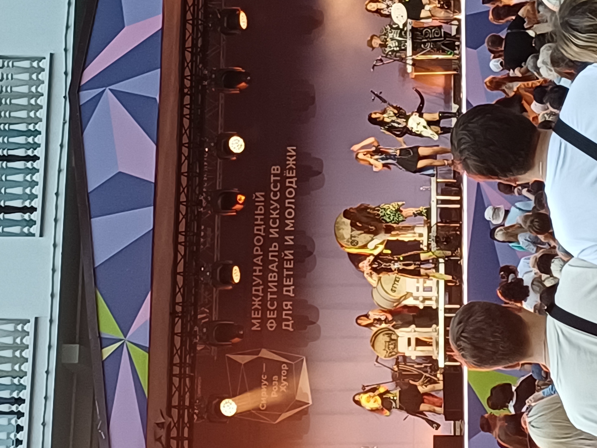 This image depicts an outdoor music festival stage in Estosadok, Russia. The stage features a band performing with multiple female musicians on instruments. Behind the performers is a large, dark backdrop with Russian text indicating "INTERNATIONAL FESTIVAL OF ARTS FOR CHILDREN AND YOUTH" and a logo with the text "Sirius – Rosa Khutor". Several spotlights are directed at the stage. In the foreground, the backs of several audience members are visible, suggesting the photograph was taken from within the crowd looking towards the stage. The environment appears to be set up for a large public event, likely in the evening or late afternoon given the lighting conditions.