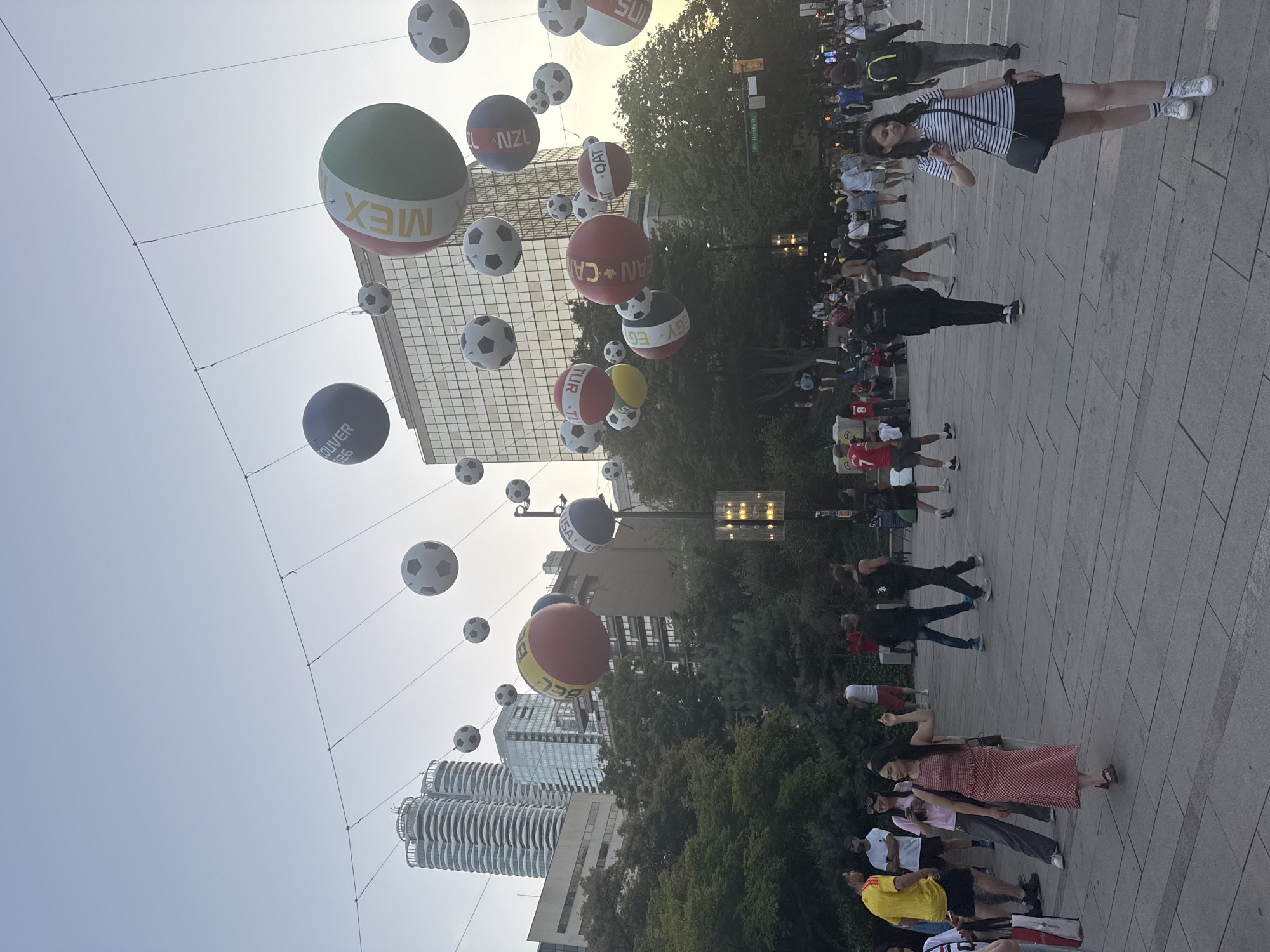 The image depicts a street scene in Vancouver, Canada, likely in a downtown area, judging by the modern high-rise buildings in the background. The sky is a pale, overcast blue, suggesting it is either early morning or late afternoon. Overhead, a series of large, inflatable balloons designed to resemble soccer balls are suspended by wires. Some of these balloons are plain black and white, while others are decorated with the flags or names of various countries, including Mexico, New Zealand, Turkey, and Egypt.
Below the balloons, a public plaza paved with large, grey stone tiles is visible. The plaza is populated by a diverse group of people, most of whom appear to be casually dressed. Many are walking or standing, some in small groups. In the foreground on the right, a young woman in a striped t-shirt and black skirt is walking away from the camera, looking back over her shoulder. To the left, a woman in a red and white checkered dress is walking forward, gesturing with her right hand. Other individuals are scattered throughout the plaza, some with their backs to the camera, others facing it.
Behind the plaza, lush green trees form a dense canopy, suggesting a park or a street lined with mature trees. Further in the background, the distinct architecture of modern skyscrapers rises above the trees. One set of buildings features a unique, undulating facade, while others are more rectilinear. A street sign is partially visible, reading "Hornby St.", confirming the location as Vancouver. The overall atmosphere appears to be relaxed and public, possibly during an event or a gathering related to a sporting occasion.