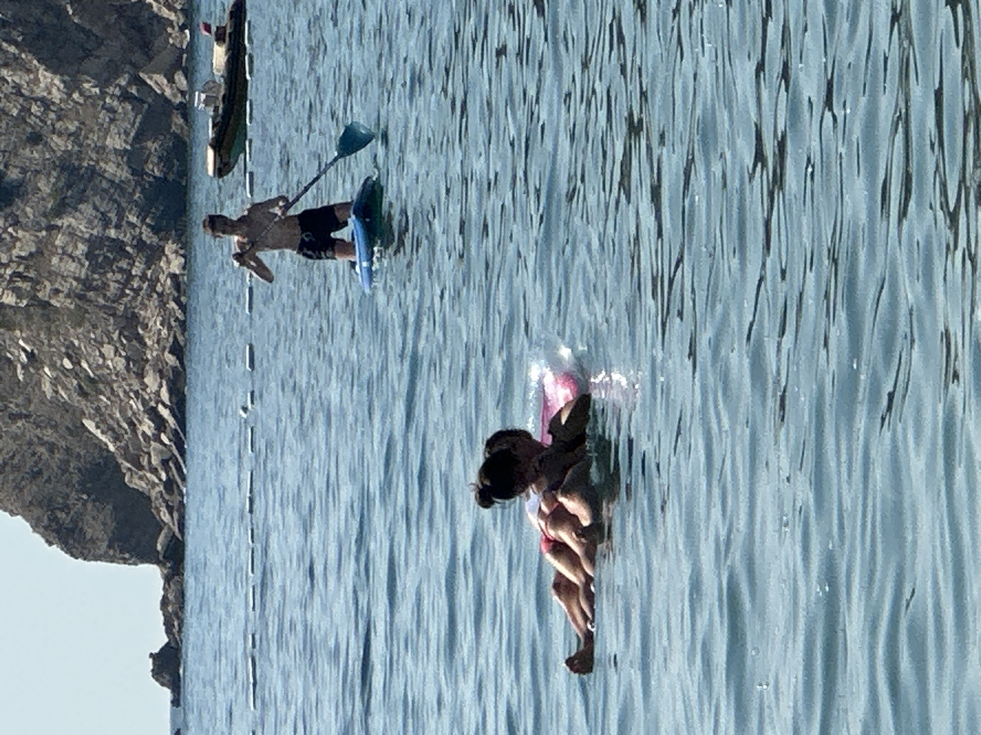 Two individuals are in the water near the shore. One person is on a paddleboard, standing and holding a paddle. In the foreground, two individuals are lying on a pink float. In the background, there is a rocky cliff, a body of water, and a boat. The scene appears to be in a coastal area, possibly Đurovići, Montenegro, given the rocky cliffs and clear blue water.