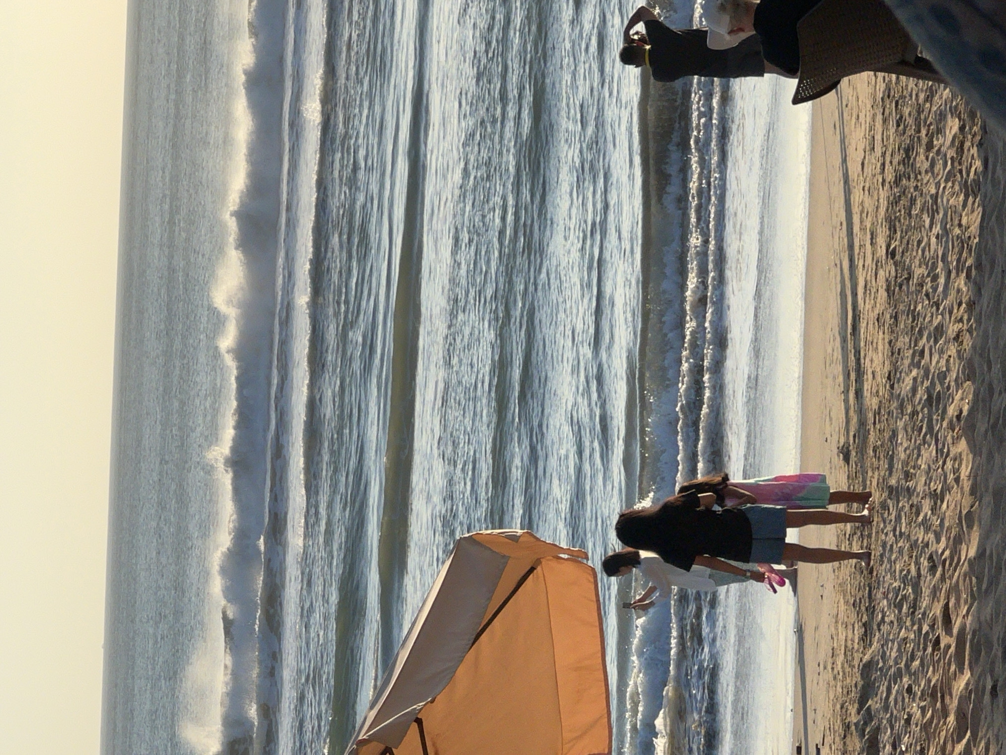 The image depicts a beach scene in Kuta, Indonesia, with the ocean and sandy shore as the primary elements. Several people are present. In the foreground, a large orange and beige beach umbrella shades a section of the sand. To the left of the umbrella, a woman in a white shirt and light-colored pants is seen holding a phone, seemingly taking a photo. Beside her, a woman in a black t-shirt and a denim skirt stands with her back to the camera, holding pink flip-flops. A young girl in a colorful dress stands next to the woman in the black t-shirt. In the middle ground, a man in a black shirt and shorts is standing near the water, holding a camera to his face. Further to the right, another person wearing a hat is seated on a chair, their back to the viewer. The ocean features rolling waves and whitecaps, indicative of active surf conditions. The sky is bright and hazy, suggesting it is either early morning or late afternoon. The sand is textured and appears dry in the foreground, with some shadows cast by the umbrella and people.