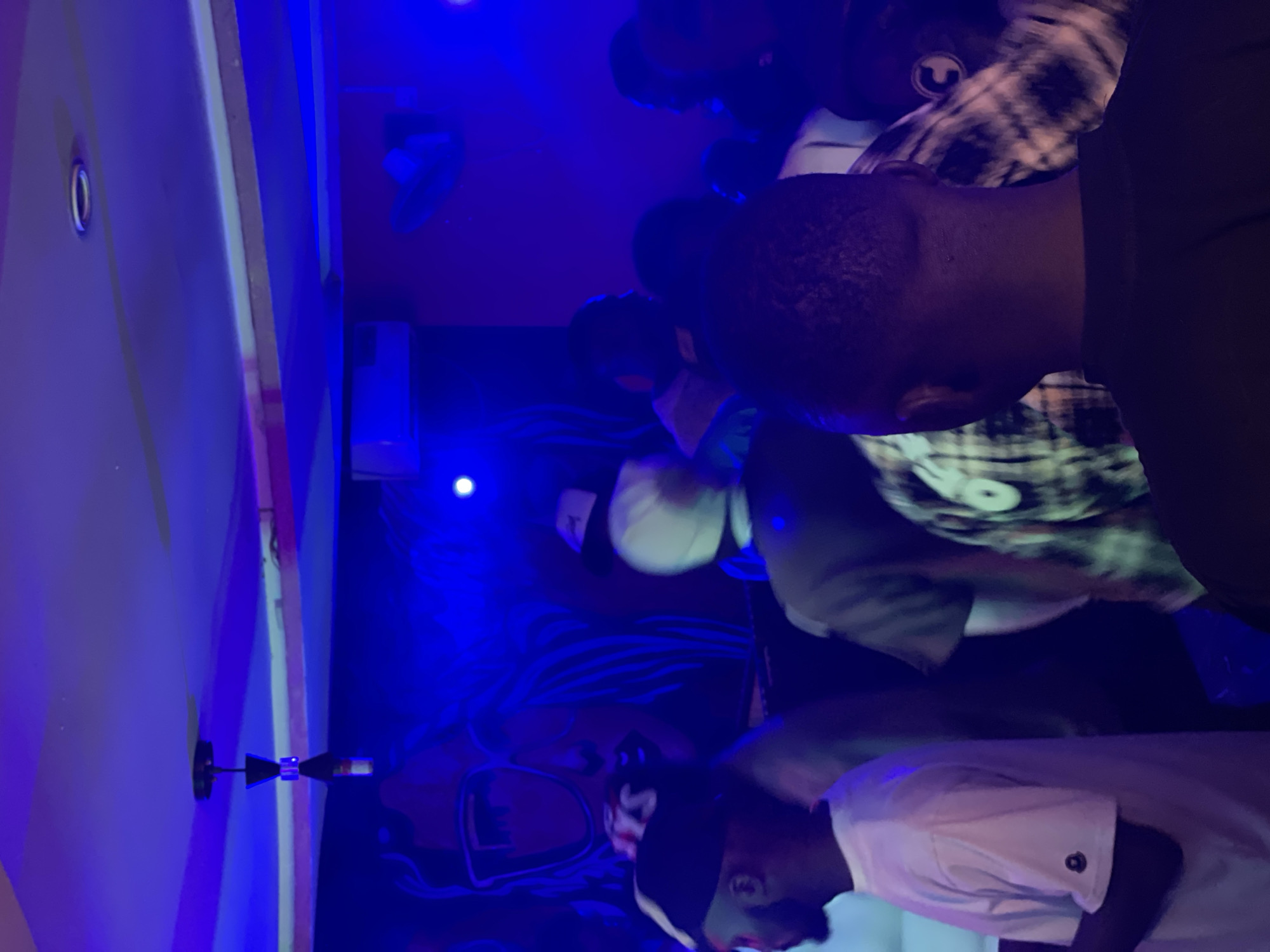 The image depicts a dimly lit indoor gathering, likely a club or lounge, with dominant blue lighting. In the background, a stylized mural of a woman with large sunglasses is visible. Several individuals are present, mostly seen from the back or side, suggesting a social event. One person in the foreground is wearing a white t-shirt with a small circular emblem on the sleeve. Another individual in the mid-ground wears a plaid shirt. The overall atmosphere appears casual and social. The specific location in Durumi, Nigeria, is not identifiable from visual cues within the image itself.