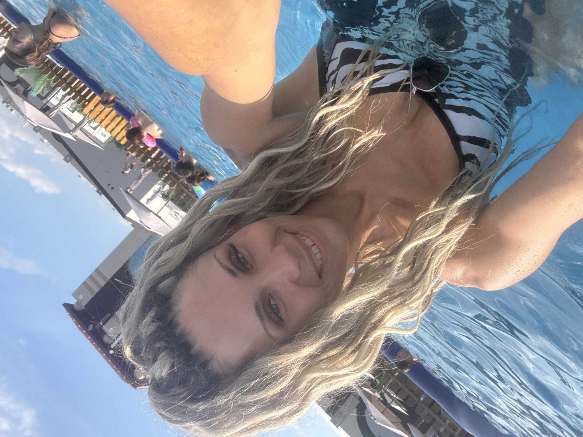A woman with blonde hair styled in dreadlocks is taking a selfie in a swimming pool. She is wearing a black and white striped swimsuit and sunglasses. Several other people are visible in the background, some of whom are also in the water. The pool area appears to be outdoors, with buildings and umbrellas visible in the distance. The sky is clear and blue. The location is the city of Rusanovo, Russia.