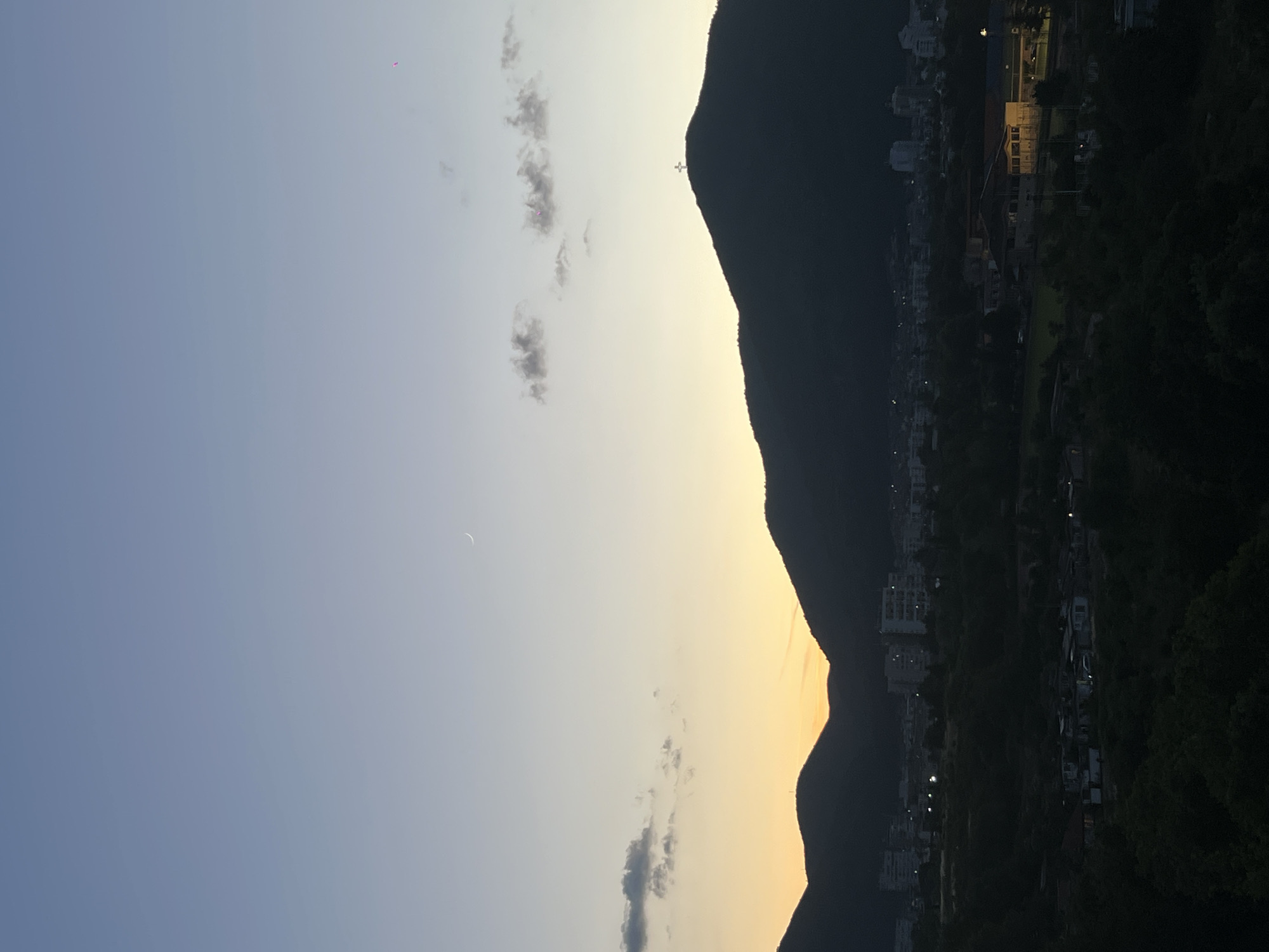 This image depicts a twilight view of the city of Sliven, Bulgaria, with the Stara Planina mountain range serving as a dramatic backdrop. The sky transitions from a soft twilight blue at the top to a warm orange glow near the horizon, indicating either sunrise or sunset. A crescent moon is visible in the upper portion of the sky. A prominent cross is illuminated atop one of the dark, silhouetted mountain peaks.
In the mid-ground, the city of Sliven is partially visible. Buildings, including several multi-story structures, are scattered across the landscape, with some lights illuminating windows and streetlights. The lower part of the image shows a foreground of dark foliage, suggesting a vantage point from a higher elevation overlooking the city. The overall atmosphere is peaceful and serene, capturing the transition from day to night.