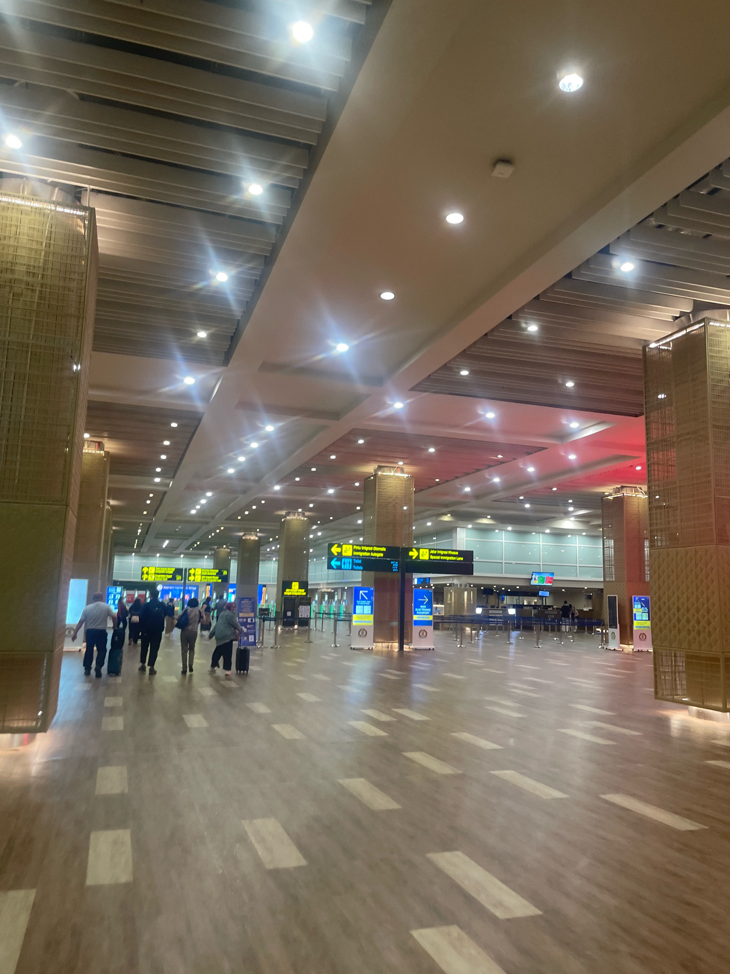 The image shows a wide, well-lit interior hallway within an airport terminal. The ceiling is high and features a modern design with rows of light fixtures. Large, square, light-colored pillars are spaced throughout the hall, some adorned with decorative, textured panels. The floor is a polished, wood-toned material with a pattern of lighter rectangular inlays.
In the mid-ground, several people are walking away from the camera, some pulling luggage. They are dressed in casual attire. Further down the hall, there are immigration counters visible, with barriers and signs. Yellow directional signs with text in what appears to be Indonesian and English indicate immigration and special immigration lanes. Some areas of the hall are illuminated with red lighting. The overall environment suggests an active airport during operational hours. This scene is consistent with the interior of Ngurah Rai International Airport in Kuta, Indonesia, a major international gateway to Bali.