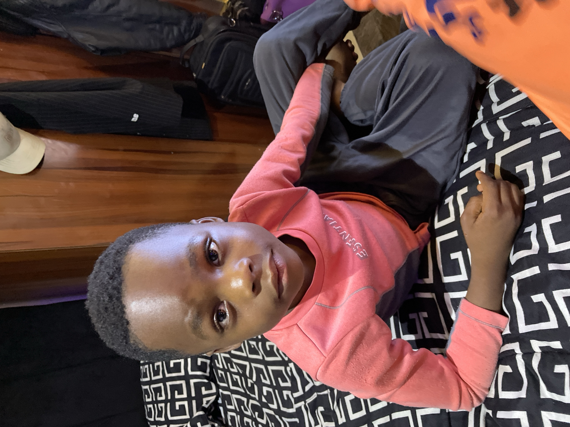 A boy is lying on a bed with a black and white geometric patterned sheet. He is wearing a pink sweatshirt and gray pants. To the left of the bed is a wooden surface with luggage on it. The photo appears to be taken indoors. The location in the city of Jos, Nigeria is not discernible from the image.