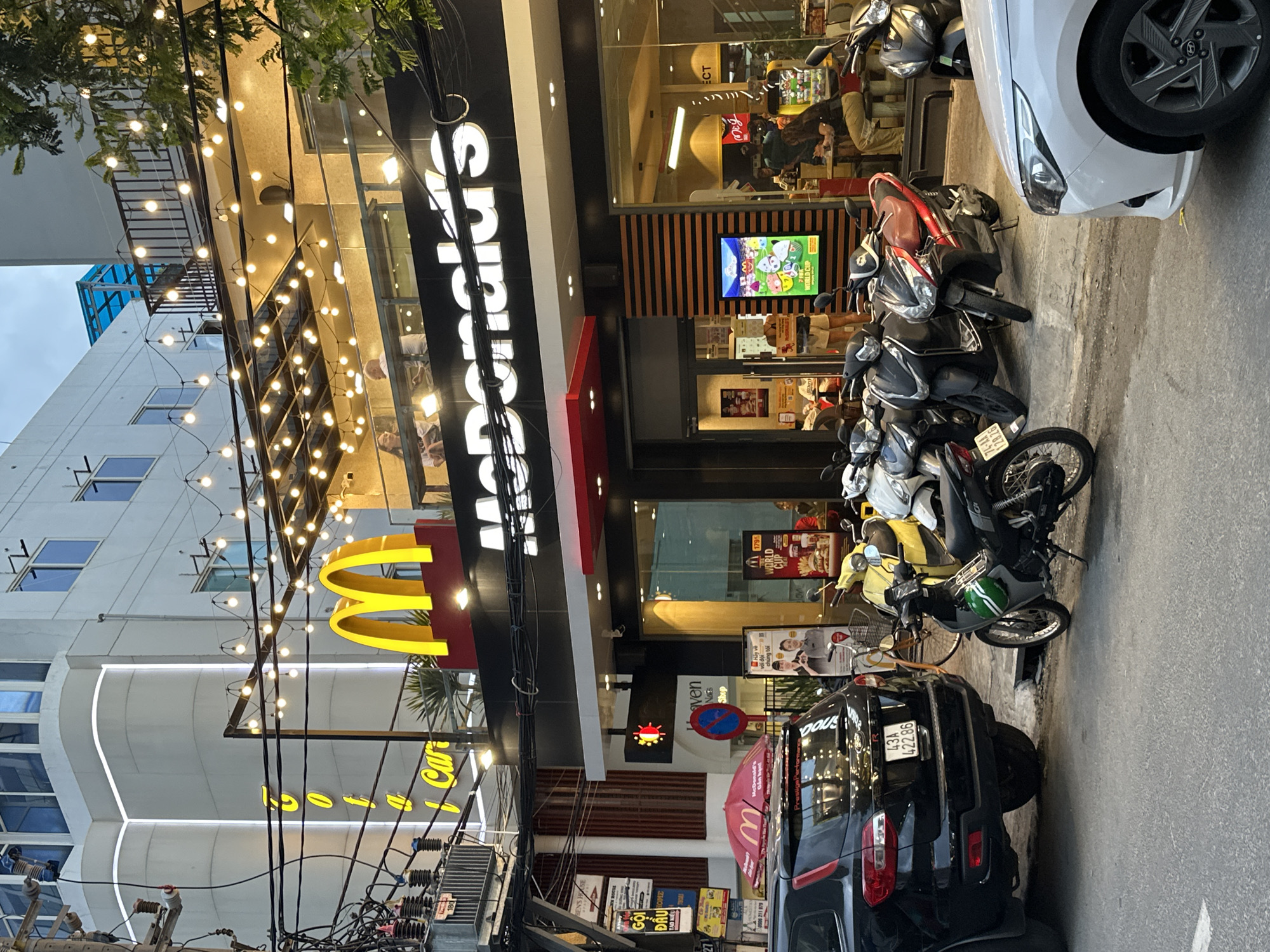 The image shows the exterior of a McDonald's restaurant on a street in the city of Đà Nẵng, Vietnam. The McDonald's signage is prominently displayed, featuring the iconic golden arches and the restaurant's name in large white letters. Strings of fairy lights are draped across the facade, illuminating the scene. Several motorbikes are parked in front of the restaurant, a common sight in Vietnam. A black SUV is also parked nearby, with its license plate clearly visible. Inside the restaurant, patrons can be seen dining. The overall scene depicts a typical urban streetscape in Vietnam, with a focus on a popular fast-food establishment.