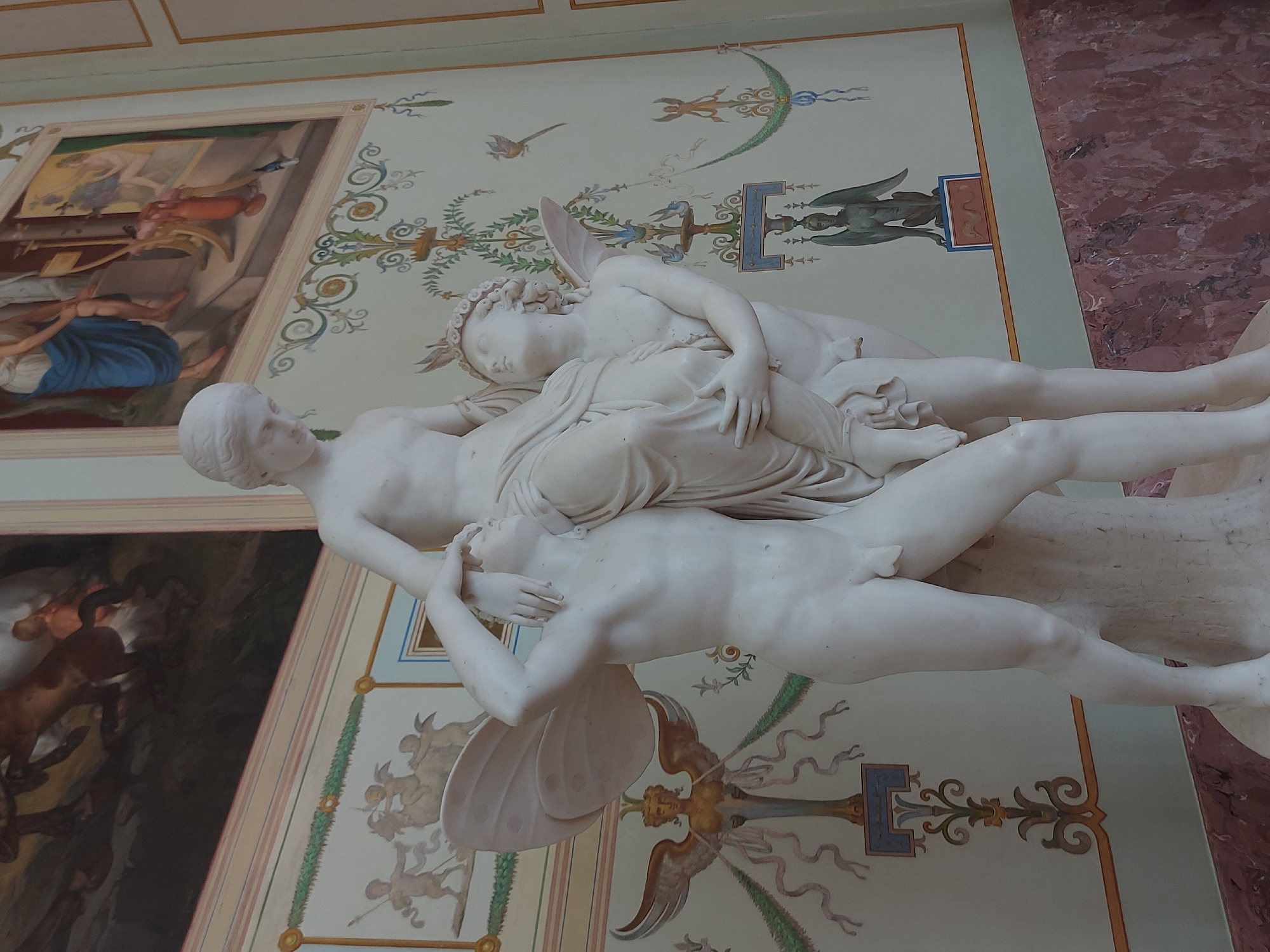 This image features a marble sculpture of Cupid and Psyche, likely a reproduction of Antonio Canova's famous work, displayed against a backdrop of ornately decorated walls. The scene is most likely located within the Hermitage Museum in Saint Petersburg, Russia, given the museum's extensive collection of classical art. The sculpture depicts the winged Cupid leaning over Psyche, their bodies intertwined. The walls behind the sculpture are adorned with detailed frescoes or murals, featuring classical motifs like floral arrangements, urns, and mythological figures. The overall impression is of a grand, historical space dedicated to art and culture.