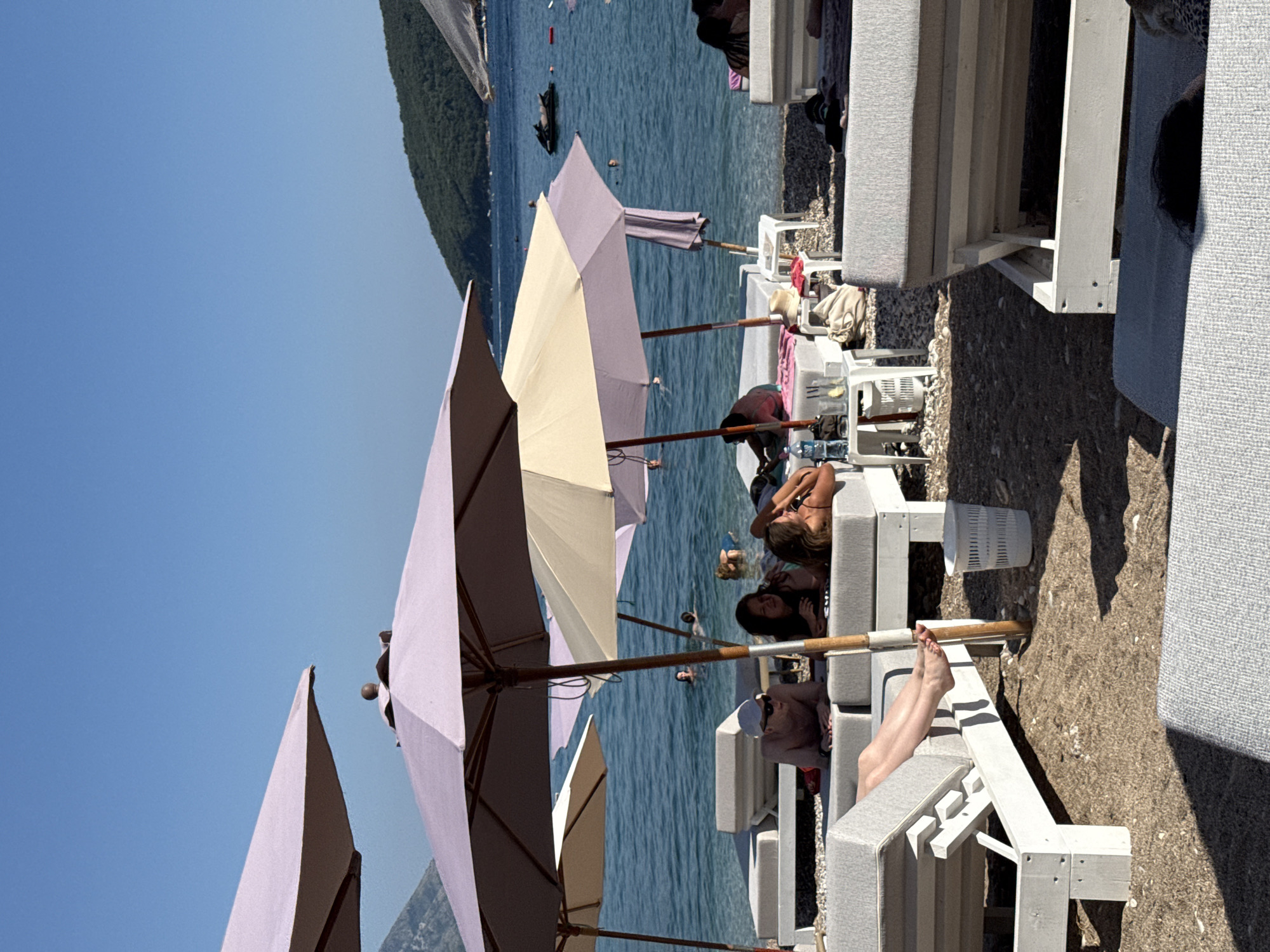 The image depicts a sunny beach scene in Budva, Montenegro, with several people relaxing under umbrellas. In the foreground, white lounge chairs with gray cushions are arranged on the pebbly beach. Some of these chairs are occupied by individuals sunbathing or lounging. In the middle ground, more lounge chairs and umbrellas are visible, with several people in the shallow, clear blue water swimming or wading. A jet ski is visible in the distance on the water. The background shows a verdant, tree-covered hillside meeting the clear blue sky. The atmosphere is relaxed and summery, indicative of a popular tourist destination.