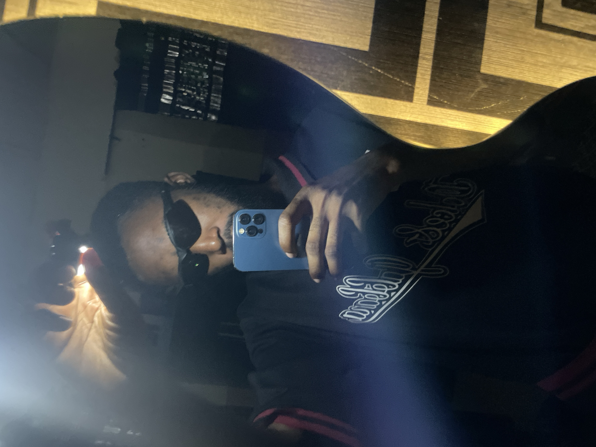 The image is a reflection in a mirror, showing a person holding a smartphone and taking a selfie. The person is wearing sunglasses and a dark t-shirt with white lettering. The background is dark and appears to be indoors, possibly a bedroom or bathroom, with a wooden cabinet visible. The image has a hazy, possibly light-leak effect, suggesting a low-light environment or a camera issue. The overall scene is personal and informal. It is not possible to determine the specific location in Lokoja, Nigeria, from the visual information provided.