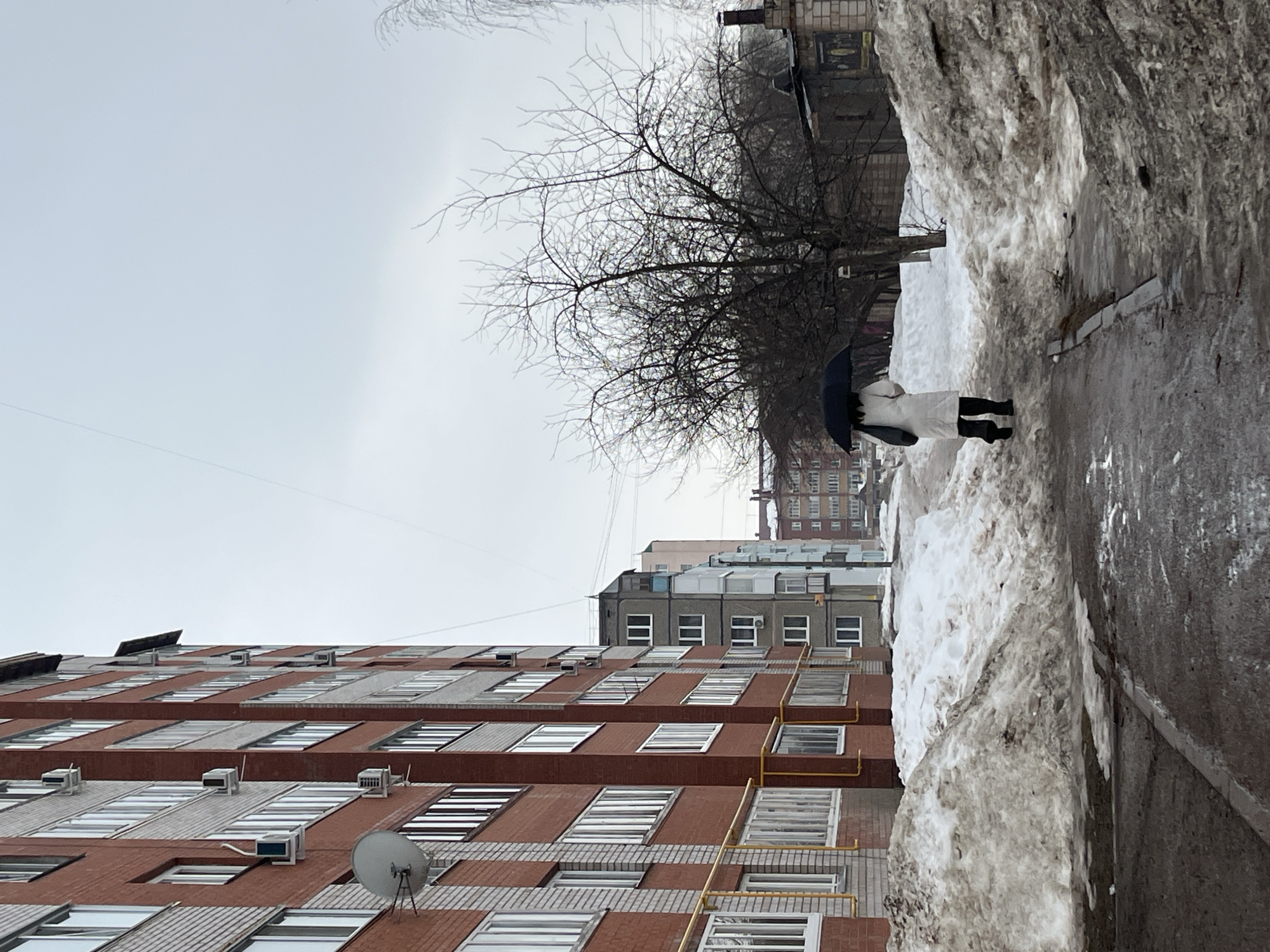 An outdoor scene in Neftekamsk, Russia, depicts a single individual walking away from the camera on a wet, paved path flanked by large, dirty snow piles. The person wears a long white coat, dark trousers, and dark footwear, holding a black umbrella and carrying a dark bag over their left shoulder. To the left, a multi-story residential building with red brick and light-colored sections features numerous windows, external air conditioning units, and yellow gas pipes. In the background, additional multi-story residential buildings of varying colors and architectural styles are visible. Bare deciduous trees, indicating winter or early spring, stand to the right of the path, partially obscuring a smaller brick building with a chimney. The sky is uniformly overcast and light grey.