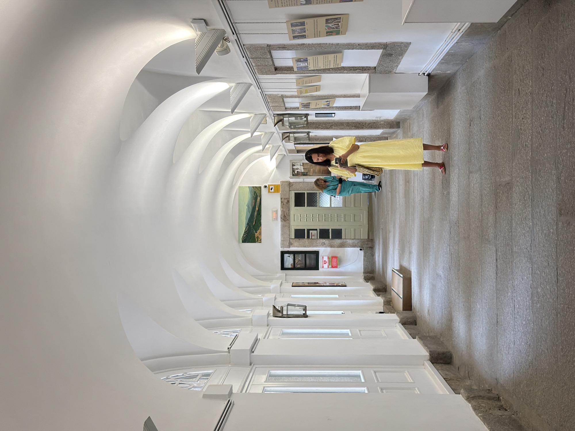 The image depicts a long, arched hallway inside a building, likely a museum or historical site. The architecture features a series of white, curved ceilings that create a cloistered effect. White walls line the left side of the hallway, punctuated by tall, white doors with glass panes. On the right, the wall is also white, with stone frames around doorways or display areas. Several framed documents or artworks are hung at intervals on the right wall, some suspended by chains.
In the foreground, a woman in a bright yellow dress stands facing the camera, holding a mobile phone in her hands and looking down at it. She is wearing pink sandals. Behind her, further down the hallway and to the left, another person, possibly a staff member, is wearing a teal uniform and appears to be looking at something on the wall. The floor is paved with large, grey flagstones. Lighting comes from modern, angled fixtures mounted on the ceiling. A landscape painting is visible on the ceiling in the distance, and a red AED (Automated External Defibrillator) box is mounted on the wall on the right. The overall impression is one of serene, institutional architecture.
This hallway appears to be located within the Royal Site of San Lorenzo de El Escorial, a UNESCO World Heritage site in San Lorenzo de El Escorial, Spain. The architectural style and the presence of artworks and historical elements are consistent with the grandeur and educational function of the Royal Site, which includes a palace, a basilica, a pantheon, a library, and a monastery.