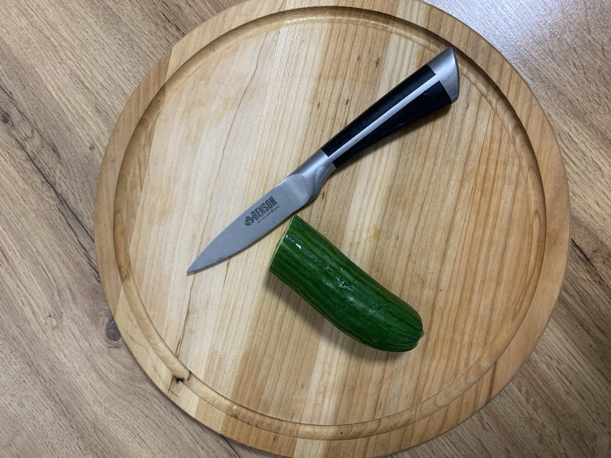 A close-up, top-down shot shows a paring knife and a partially cut cucumber resting on a round wooden cutting board. The cutting board is placed on a light-colored wooden surface, likely a table or countertop, with a distinct wood grain running horizontally.
The knife, positioned diagonally across the board, has a silver-colored blade and a handle with black and silver sections. Clearly visible on the blade is the brand name "BENSON" and the tagline "It's made your life easy." Next to the knife is a dark green cucumber, which has one end neatly sliced off, revealing its light green interior and suggesting food preparation is in progress or just completed. The cucumber's skin has visible ridges and appears fresh.
The round wooden cutting board has a light, natural wood tone and a shallow, circular groove carved near its perimeter. The overall lighting is bright and even, indicating an indoor setting, likely during the day. No people are visible, and the scene evokes a calm, domestic atmosphere. This image was captured in Ternopil, Ukraine.