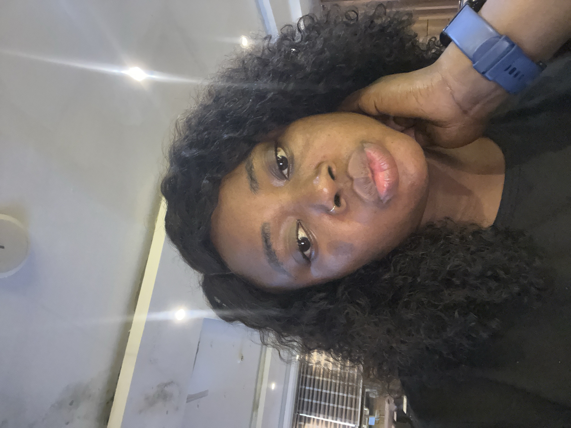 A medium shot shows a Black woman from the chest up, looking directly at the camera. She has curly black hair, brown eyes, and a nose ring. She is wearing a black t-shirt and a blue smartwatch on her left wrist. Her right hand is resting on her cheek, with her fingers extended. The background is blurred, but appears to be an indoor setting with fluorescent lighting and what looks like blinds on a window. The lighting suggests it could be daytime, possibly in an office or public space. There are no visible elements that specifically indicate the location is in Abuja, Nigeria.