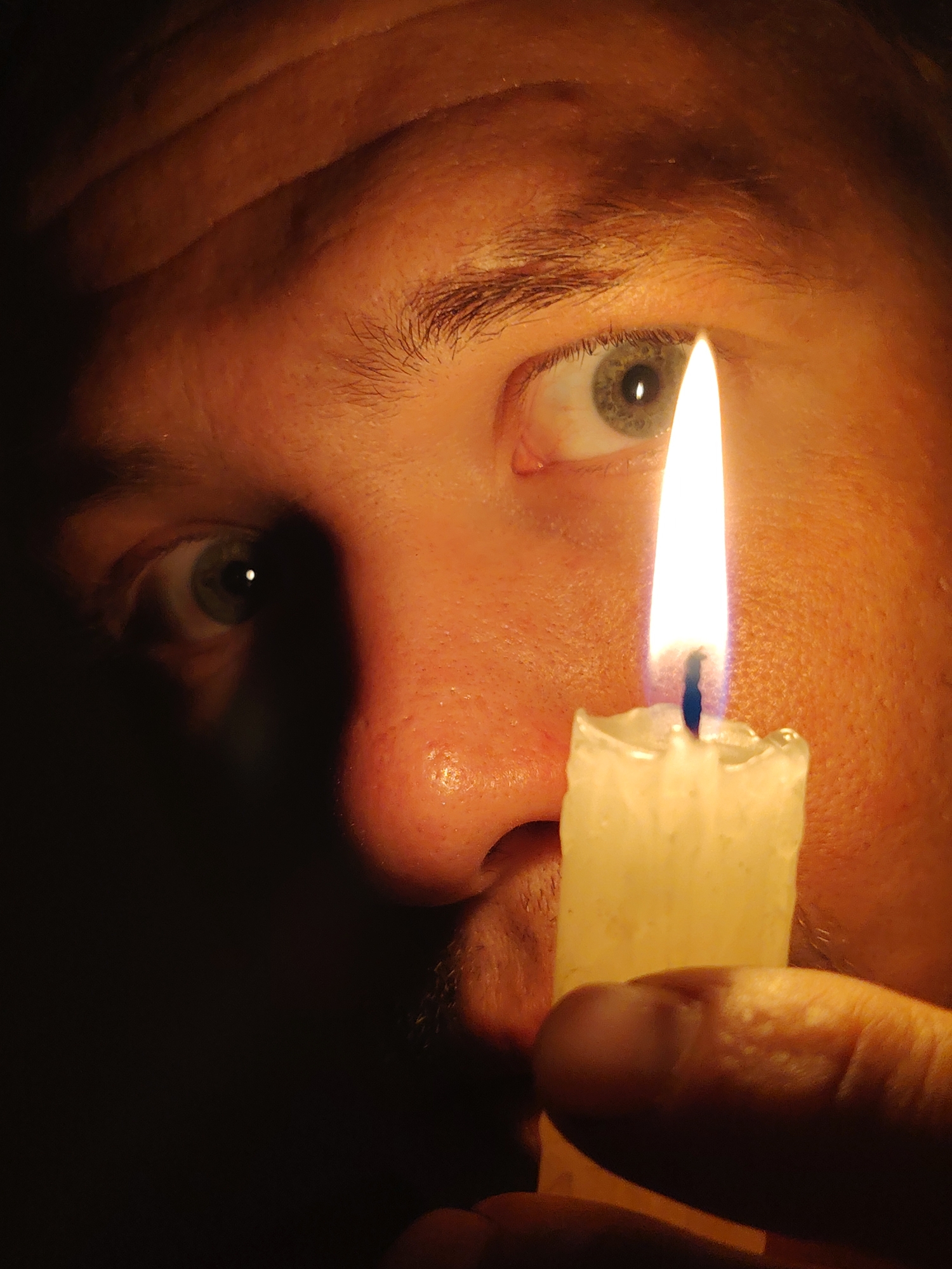 The image shows a close-up of a person's face, illuminated by a lit candle. The focus is on the eye and nose area, with the flame of the candle positioned between them, casting a warm glow. The person's skin appears textured, and the eyebrow is visible. A finger holding the candle is also in the frame, obscuring part of the lower face. The background is dark, creating a dramatic effect. There is no indication of the city of Chaikovsky, Russia in the image.