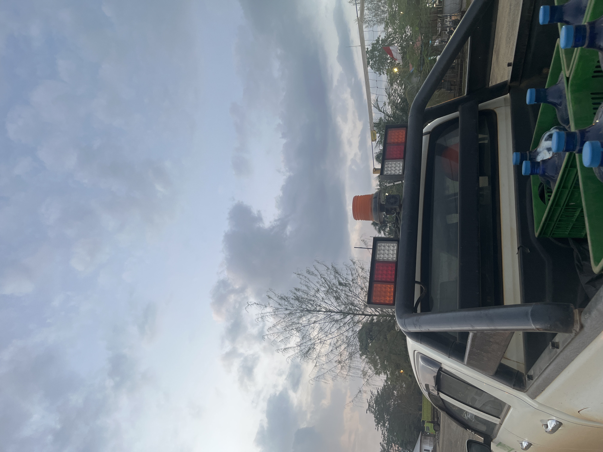 An outdoor, low-angle shot captures the rear of a white pickup truck under a cloudy sky, possibly in Unknown, Indonesia, as suggested by the presence of a red and white Indonesian flag near a volleyball net. The truck bed is filled with green crates containing multiple large blue-capped water bottles. Mounted on the truck's roll bar are two rectangular light fixtures, one with amber and red lights and the other with white lights, along with an orange rotating beacon. In the background, a tall, thin tree with sparse foliage stands out against the muted sky, and a volleyball net with a flag is visible. A green dump truck is parked in the distance. The overall atmosphere suggests a rural or semi-urban setting at dusk.