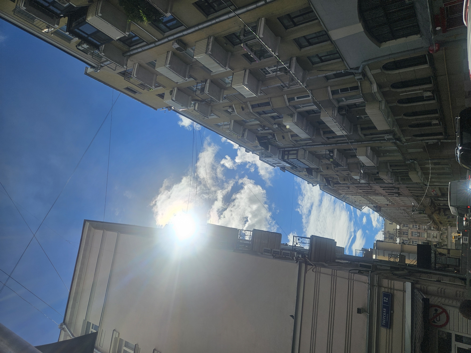 Looking up from street level in Moscow, Russia, the photograph shows an upward view of buildings and the sky. The buildings are varied in height, color, and architectural style, with some appearing to be older residential buildings with balconies and air conditioning units. Power lines crisscross the space between buildings. The sky is blue with some scattered clouds, and the sun is positioned between two buildings, causing a glare. A street sign in Russian "Тверская 12" (Tverskaya 12) is visible on one of the buildings, indicating that the location is Tverskaya Street. A portion of traffic, including a car, is partially visible on the right side of the image.