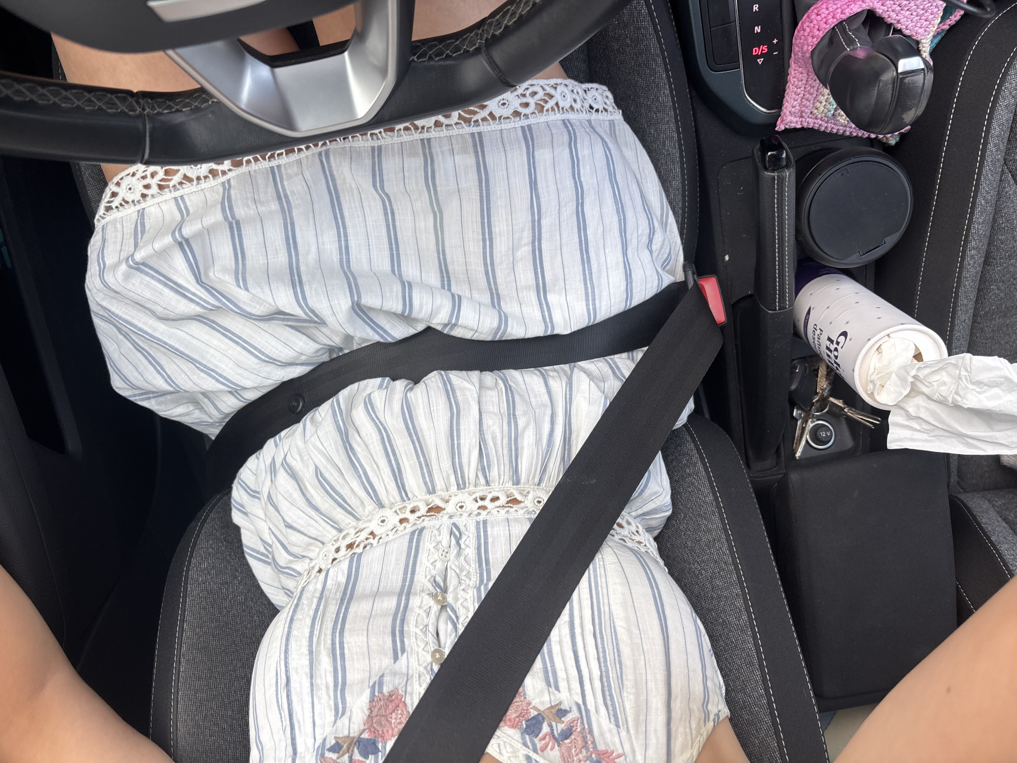 A person is seated in the driver's seat of a car, wearing a white and blue striped off-the-shoulder top with floral embroidery, secured by a black seatbelt. The steering wheel is visible in the upper left corner. The car's center console area includes a gear shift, a cup holder with a paper cup, keys, tissues, and headphones covered in a pink and purple crochet holder. The image was taken in a vehicle, the location in the city of San Andrés Cholula, Mexico is not determinable from the image.