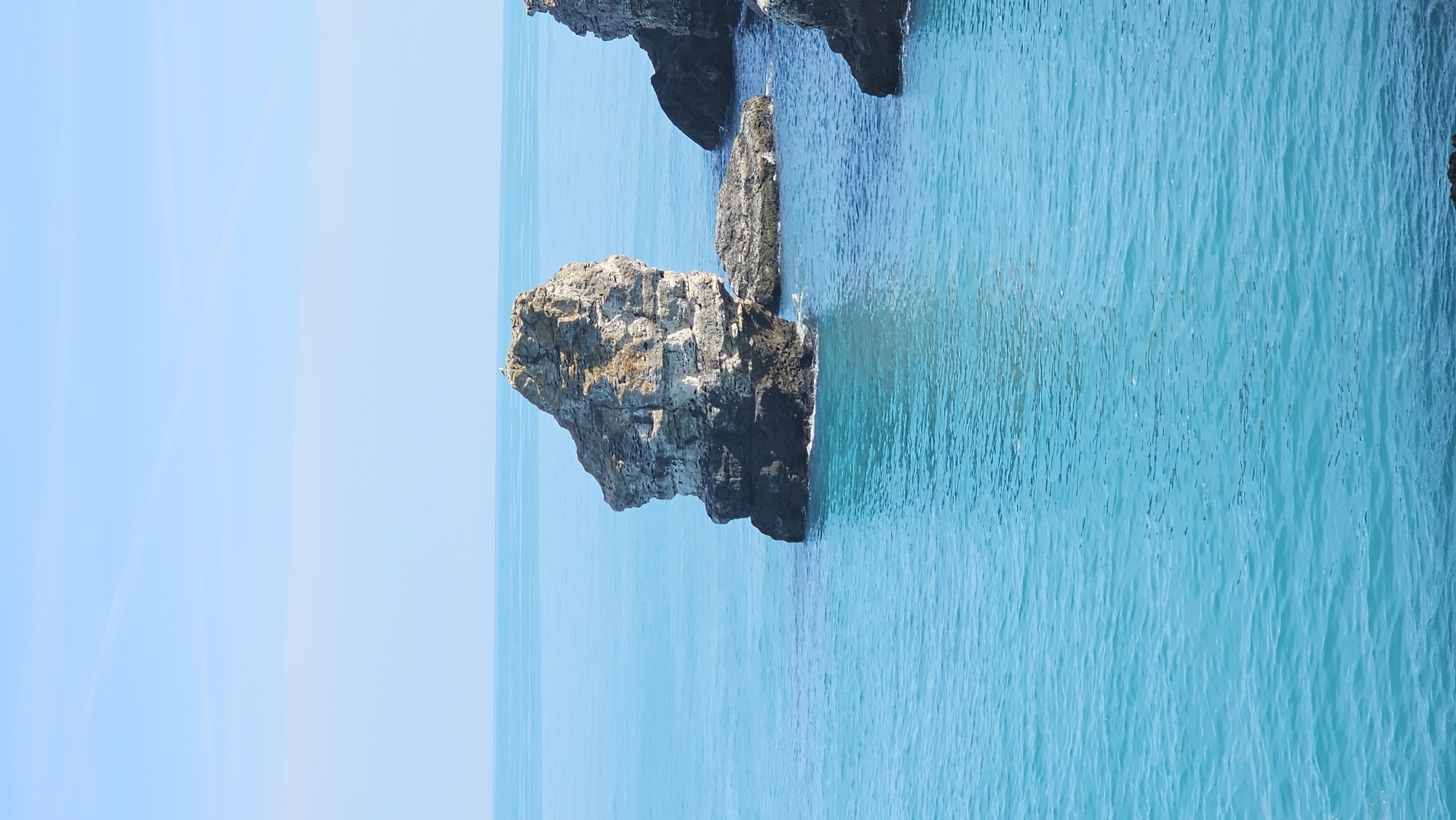 The image depicts a seascape with rock formations emerging from the turquoise water. The sky is light blue, with faint contrails visible. There are no people present in the image. The location is likely Tyulenovo, Bulgaria, known for its rocky coastline.