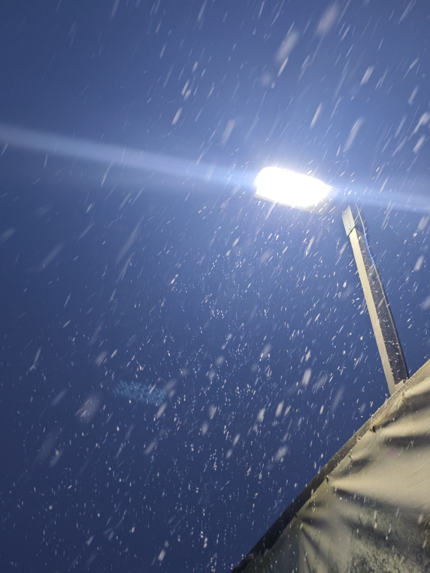 The image captures a nighttime scene during heavy snowfall, viewed from a low angle looking upwards. A bright, rectangular street light, possibly an LED fixture, is prominently featured in the upper right quadrant, casting a strong white light that illuminates numerous falling snowflakes. These snowflakes appear as distinct white streaks and dots against the deep blue, dark sky, with some showing motion blur, indicating their descent. A pronounced horizontal lens flare extends from the light across the blue background.
The street light is mounted on a slender, vertical pole, partially visible on the right side of the frame. In the bottom right corner, a portion of a light-colored, textured structure, possibly a tent, canopy, or awning, is visible, angling upwards and providing a foreground element. The scene conveys a cold, wintery atmosphere under artificial light. No people or discernible text are present in the image. The location is not identifiable from the visual content.
