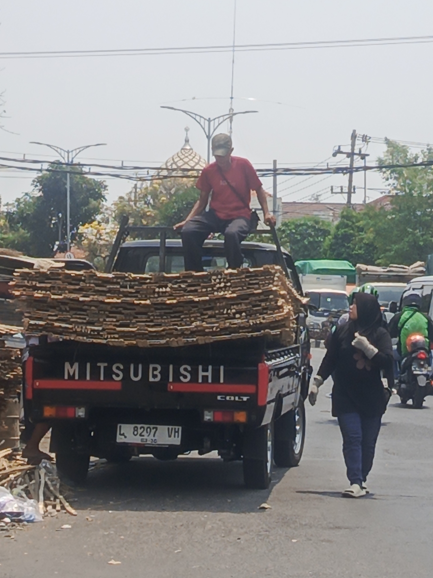 The image captures a street scene in Surabaya, Indonesia, under a bright, hazy sky. A black Mitsubishi Colt pickup truck is prominently featured, heavily laden with stacks of lumber. A man wearing a red t-shirt and a cap is perched on the edge of the truck bed, his legs dangling. In the foreground, a woman wearing a black hijab, long-sleeved black shirt, blue jeans, and white slip-on shoes is walking away from the truck. She is wearing white gloves. Behind the truck, other vehicles, including a motorcycle with a rider in a green helmet, are visible, suggesting a traffic environment. Overhead, power lines and streetlights are strung across the sky. In the background, trees and buildings, including a structure with a domed roof, contribute to the urban setting. The road appears to be asphalt, with some debris and discarded materials along the side.