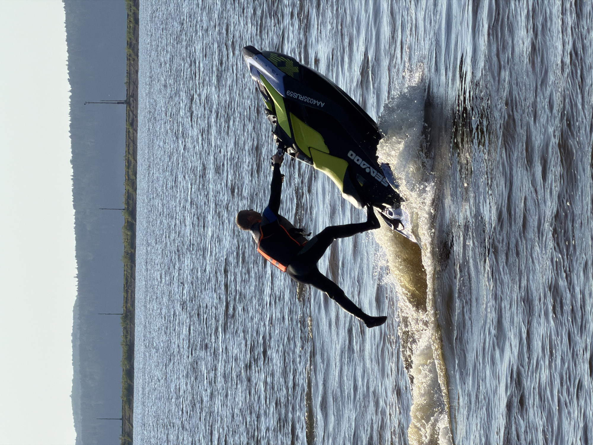 A man in a wetsuit is seen riding a Sea-Doo watercraft on a body of water. The watercraft is predominantly black with yellow and blue accents, and has the registration number AA4035RUS59 printed on its side. The man is in mid-air, appearing to be performing a trick or having been ejected from the watercraft. The water is choppy, indicating movement and speed.
In the background, the shoreline is visible, with trees and vegetation indicating a natural or semi-natural environment. The sky is overcast, suggesting subdued lighting conditions.
Based on the registration number, the location is likely in Russia, specifically in the Perm Krai region, where the vehicle registration code "59" is assigned. The city of Chaikovsky is located in this region.