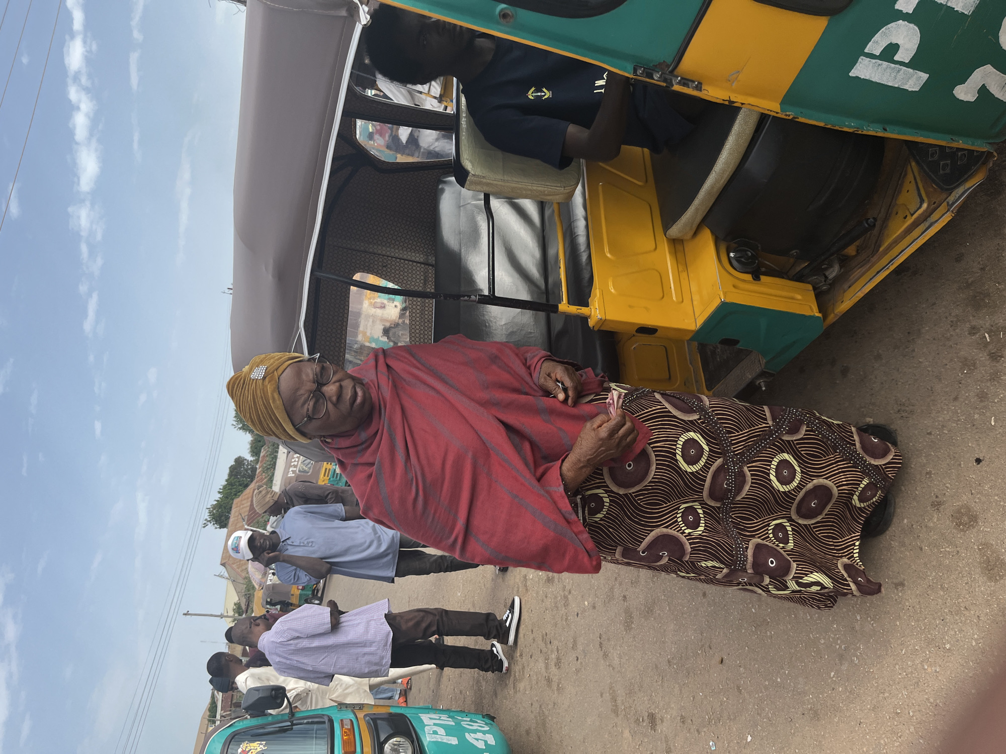 An elderly Black woman wearing a yellow headscarf and glasses stands in front of a yellow and green auto-rickshaw on a dusty road in Gura Top, Nigeria. She is wearing a red shawl draped over a brown and tan patterned skirt. Her hands are holding money. A young Black man wearing a dark blue shirt is sitting in the driver's seat of the auto-rickshaw. Several other people, including men wearing a white baseball cap and a light blue shirt, are standing in the background. The sky is partly cloudy.