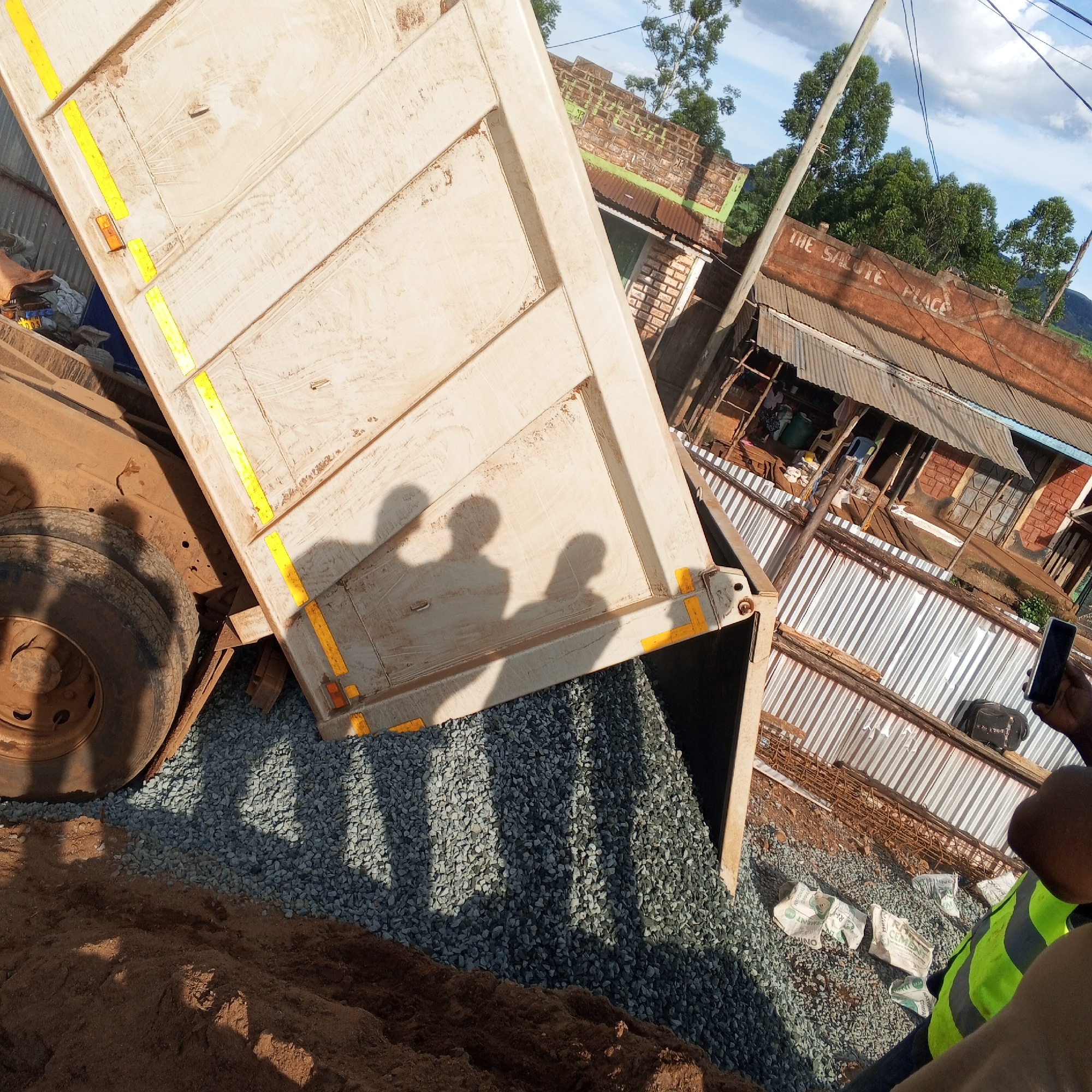 In Borabu Chitago ward, Kenya, a dump truck is tilted up, unloading a load of gravel. Two people stand near the back of the truck, casting shadows on the truck bed and the gravel. A person in a neon green vest stands at the right of the image, holding a cell phone and apparently taking a picture. A building with a sign that reads "THE SALUTE PLACE" is in the background, with power lines visible above. Some bags, rebar, and sand are present alongside the gravel.
