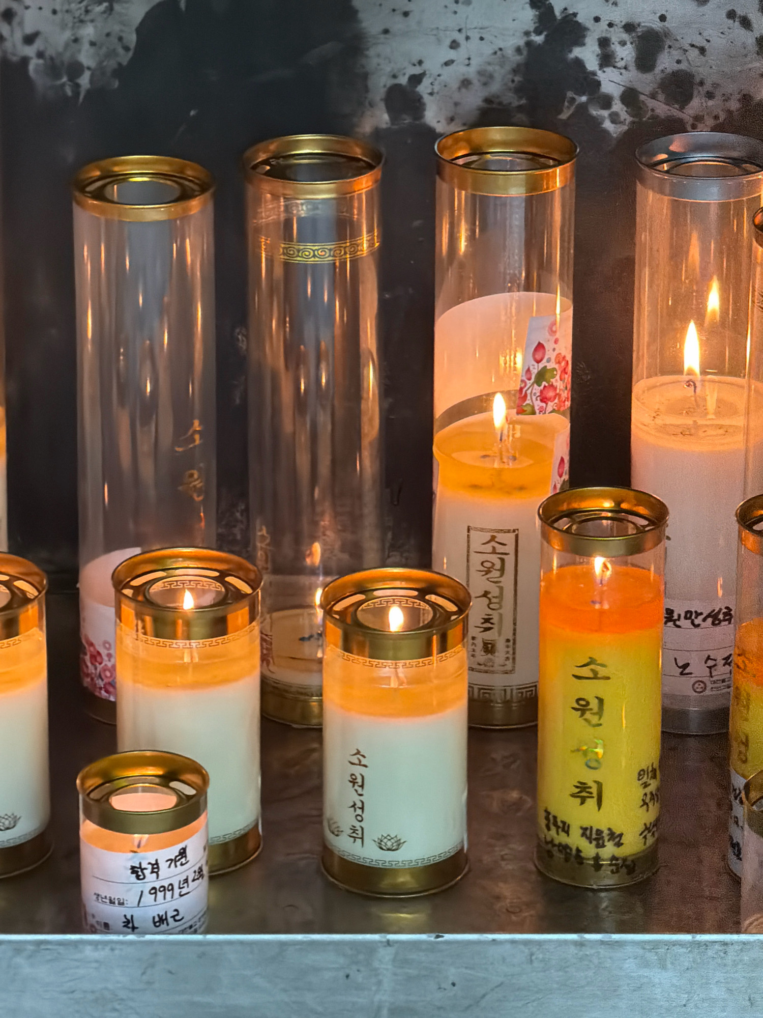 The image shows a close-up of several tall, cylindrical candles with brass tops, burning in what appears to be a temple or shrine. The candles are arranged in a row on a metallic surface, and the background is dark and textured, possibly a wall or shrine backdrop. Some of the candles have labels with Korean writing. One prominent label on a yellow and white candle reads "소원 성취" (so-won seong-chwi), meaning "wish fulfillment." Another candle has a label with a date, "1999년 2월" (1999 Nyeon 2 Wol), meaning "February 1999." The overall impression is one of reverence and dedication. The scene is located in Dongducheon-si, South Korea.