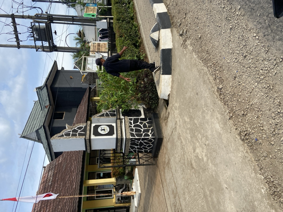 The image shows a man walking along a street in Sukatani, Indonesia (latitude: -6.608, longitude: 107.414). He is wearing a black shirt, black pants, and black shoes. He is walking to the left of the image, away from a black and white gate and a black and white pillar with a sign that reads "Welcome" in Indonesian. The gate is open and there is a small, black motorcycle parked in the background. The man is walking on a paved sidewalk that is edged by a concrete curb painted black and white. The sidewalk leads to a gravel road. There is a black and white sign on a pole in the background, and a large electrical transformer mounted on a pole with wires running to the top of the frame. There is a row of green bushes on the right side of the image. There is a tall, white building in the background, and a smaller building in the background to the left of the image, which has a red flag with a white star on the pole. The image was taken during the day, with a clear blue sky and puffy white clouds.