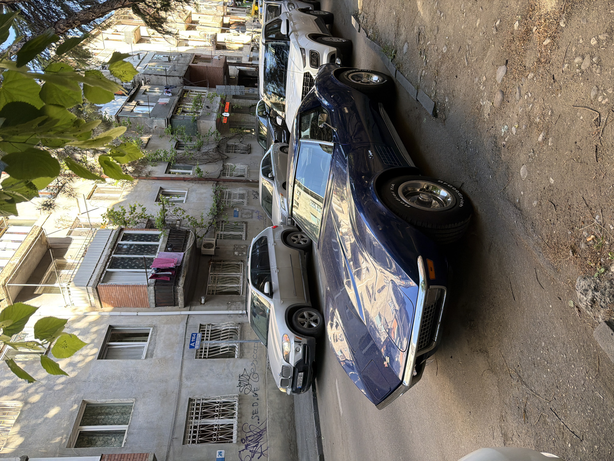 The image shows a street scene in Tbilisi, Georgia, featuring several parked cars alongside an aging building. At the forefront is a blue vintage car with a chrome bumper and visible BFGoodrich Radial T/A tires. Behind it are parked a silver SUV, possibly a BMW X5, and a white crossover SUV. Further down the line, other vehicles are partially visible.
The building on the left is characterized by its weathered facade, multiple windows, and metal window grates. Laundry hangs out to dry on a balcony. Graffiti is visible on the building's lower section. A street sign labeled "A 1164" is attached to the building.
The street itself is narrow and appears to be made of asphalt and gravel. Leaves from a tree overhang the scene in the upper left corner. The overall lighting suggests it is daytime.