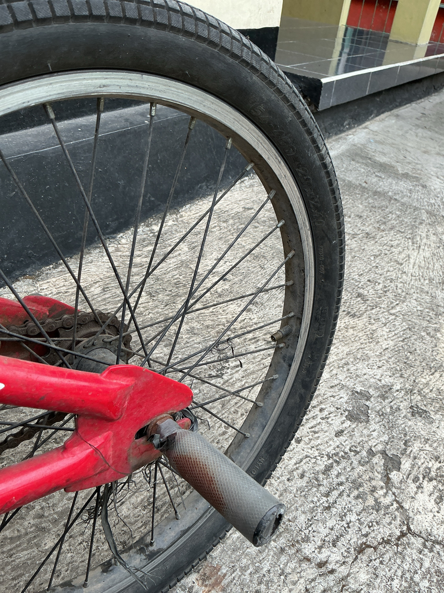 The image displays a close-up, low-angle shot of the rear wheel and frame of a red bicycle. The bicycle is positioned on a concrete surface, typical of a sidewalk or road in an urban environment. In the background, a building with tiled walls in yellow and red is visible. The bicycle's tire is worn, and the spokes of the wheel are visible, some appearing slightly bent or rusted. The red frame of the bicycle shows signs of wear and tear, with some parts appearing to be taped together. A grey, textured pedal extender is attached to the frame near the rear axle. The scene appears to be outdoors, likely in the city of Magelang, Indonesia, based on the request. There are no people or animals present in the frame. The overall impression is one of a utilitarian, well-used bicycle in an urban setting.