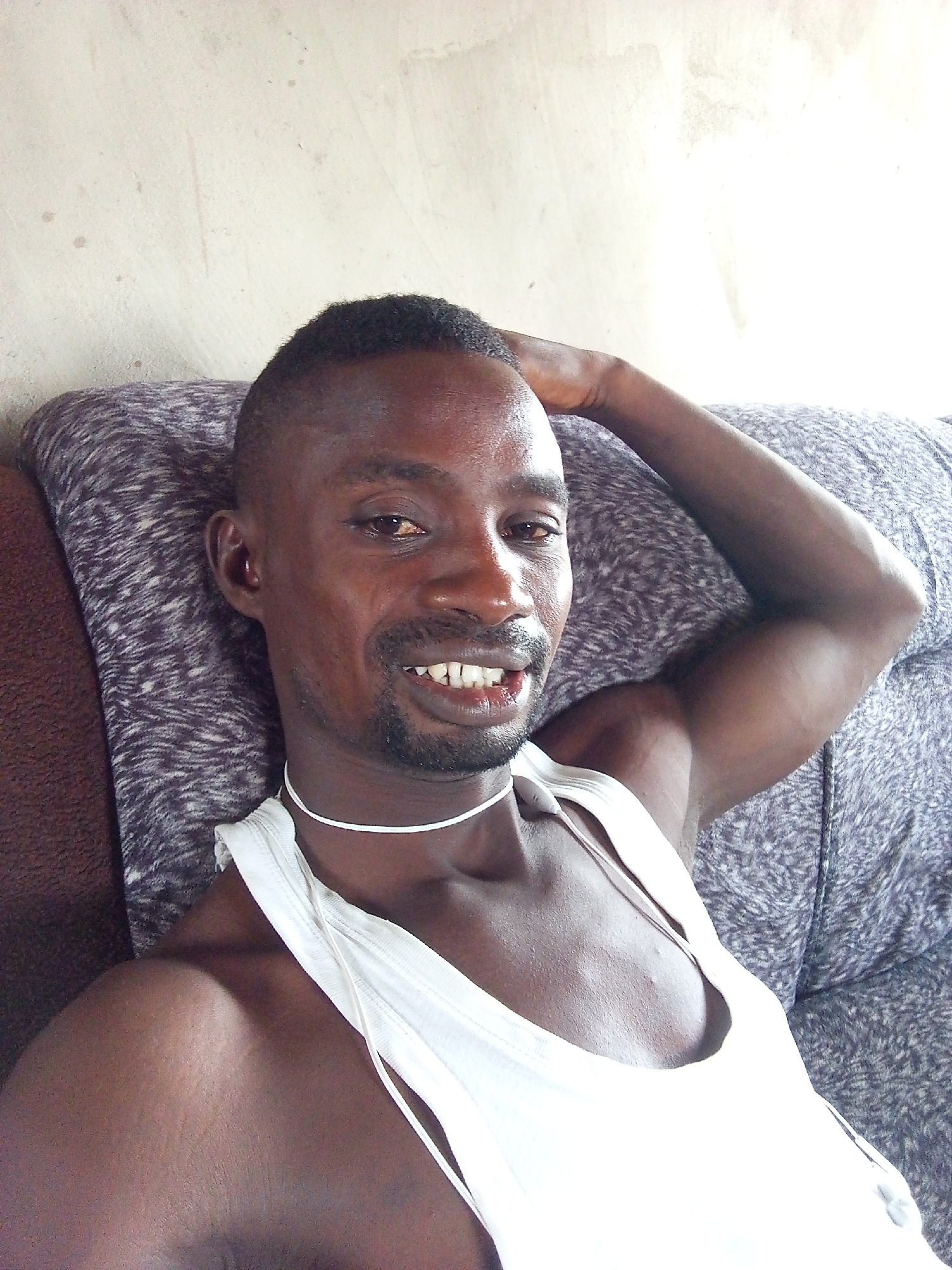 A dark-skinned man with short dark hair and a short beard is depicted in a medium close-up, smiling broadly directly at the camera. He is wearing a white sleeveless tank top, revealing his chest and arms, and a thin white cord is visible around his neck. His left arm is bent, with his hand resting behind his head, as he reclines against a patterned cushion or sofa backrest. His expression is cheerful and relaxed, with a wide smile showing his teeth.
The setting is indoors, featuring a light-colored wall in the background with some texture and minor marks. The man is seated on what appears to be a sofa, with a grey and purple patterned cushion behind him and a darker brown section of furniture visible to his left. The lighting is bright, suggesting natural light. The man appears to be in a comfortable and casual pose. No visible text is present in the image. The scene conveys a friendly and relaxed atmosphere. The location is Adonkolo, Nigeria.