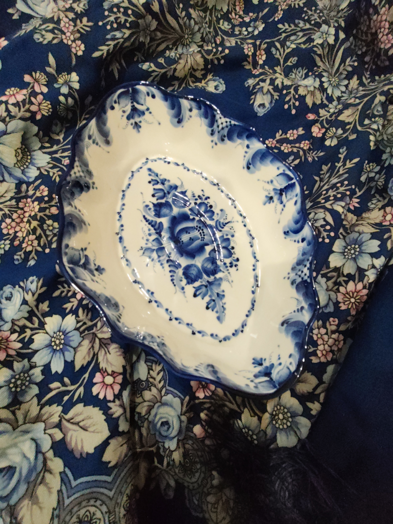 The image shows a white ceramic dish with blue floral Gzhel painting, placed on a dark blue fabric with a floral pattern. The dish is oval-shaped with a scalloped edge and a decorative border of blue brushstrokes. The floral pattern on the dish features various blue flowers and leaves. The fabric beneath has a complex floral design with blue, pink, and green tones. The scene is a still life, with no people or actions depicted. The image does not contain any information to determine its location in the city of Sochi, Russia.