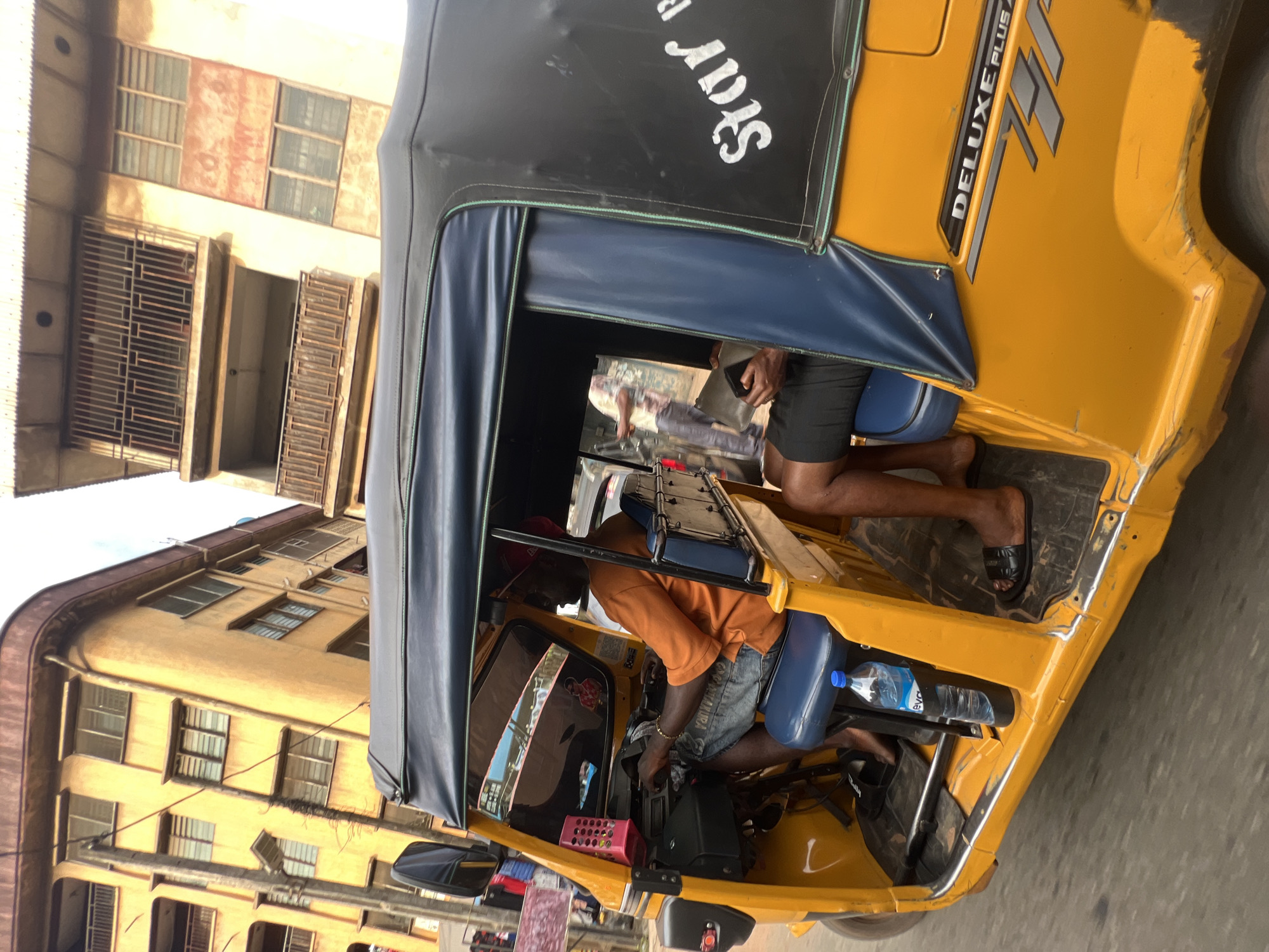 The image shows a man driving a yellow auto-rickshaw in Nkpor, Nigeria. The man is wearing an orange t-shirt and denim shorts, with a red cap on his head. He is holding the steering wheel and appears to be focused on the road. Inside the auto-rickshaw, there is a blue seat, a water bottle, and a pink basket. The auto-rickshaw has a black canopy with white lettering that reads "Star". There is also text on the side of the auto-rickshaw that says "DELUXE PLUS". The background of the image shows buildings with windows and balconies, typical of an urban setting in Nigeria. The lighting suggests it is daytime, and the weather appears to be clear. The scene captures a moment of daily life in Nigeria, with the auto-rickshaw serving as a common mode of transportation.
