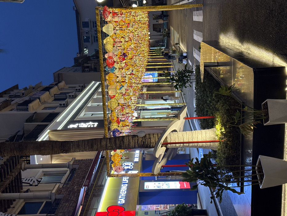 The image depicts an illuminated street scene at dusk or night in the Chongqing High-tech Zone, China. A prominent feature is an overhead canopy of numerous colorful Chinese lanterns and string lights, suspended from a golden rectangular framework spanning the street.
On the left, commercial storefronts are visible, including one with a "GATHERING LIFE" sign in English alongside Chinese characters, and another with a large red display. The architecture above these stores features stone cladding and ornate balconies. A tall palm tree trunk partially obscures the left side of the frame.
In the foreground, a dark planter bed contains green foliage and a white, abstract, multi-lobed sculpture, which is uplight by a hidden source and features vertical red accents. Adjacent to this, a long, dark bench or planter box with a reflective golden surface extends to the right, incorporating a small, round golden table. White potted plants are placed on the ground nearby.
The street surface below the lantern canopy is paved and includes white pedestrian crossing stripes. Scattered individuals are present: one person walks away on the right under the canopy, another person bends over near a planter on the right, and a third is visible further down the street. Storefronts with large glass windows and illuminated interiors line the right side of the street, with residential-style buildings and visible air conditioning units on their upper floors. The sky above is a deep indigo, indicating the nighttime setting, with artificial light sources dominating the scene.