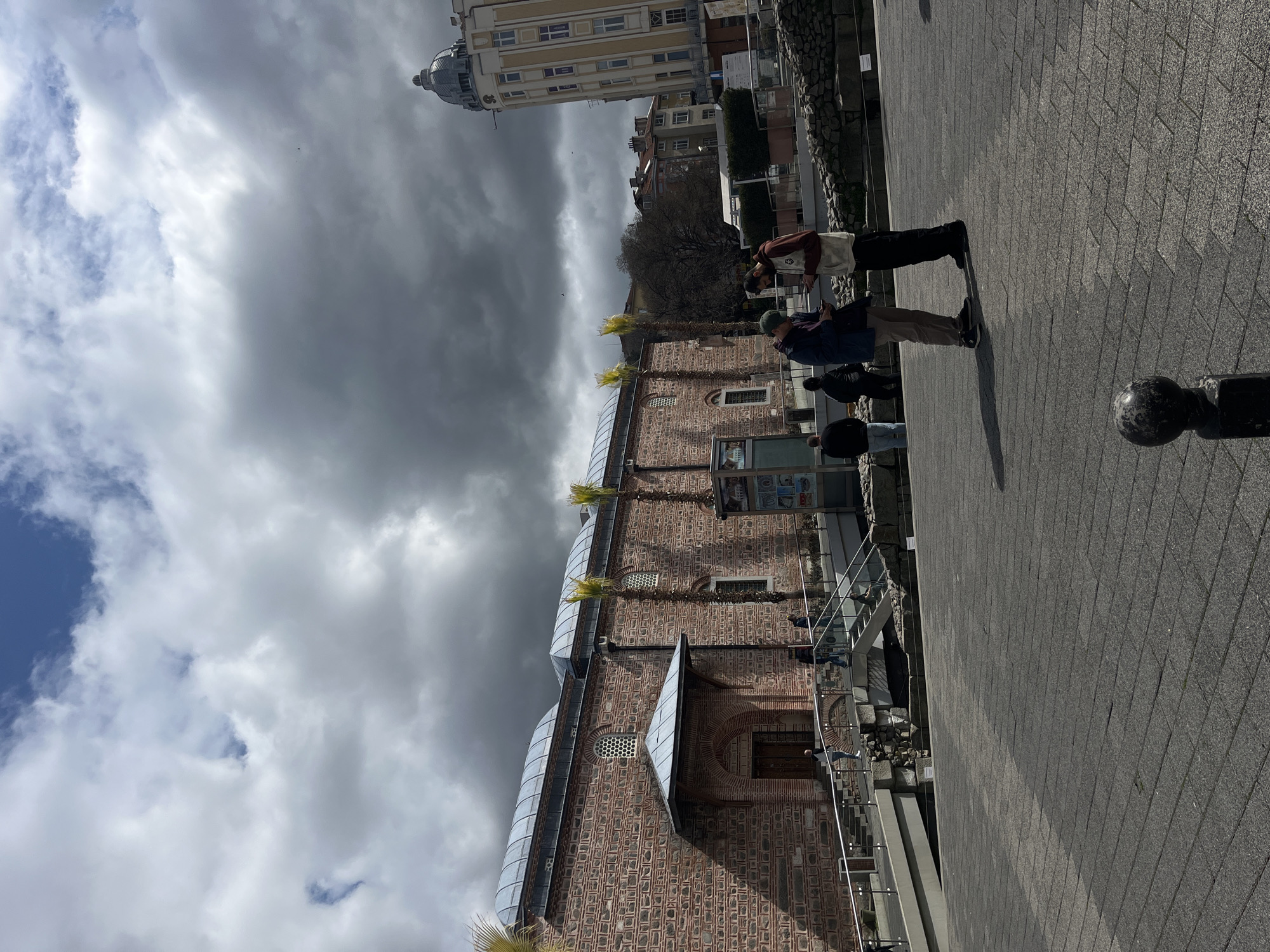 The image captures a public pedestrian area in Plovdiv, Bulgaria, situated adjacent to the Dzhumaya Mosque and archaeological remains of the Roman Stadium. The sky is predominantly overcast with dark, heavy clouds, though lighter clouds and patches of blue sky are visible in the upper left.
In the foreground, a dark grey paved plaza extends, with a spherical black bollard partially visible in the bottom center. Two males stand centrally, facing each other, both looking at smartphones. The male on the left wears a dark blue jacket, light brown pants, and a green baseball cap, holding his phone with both hands. The male on the right wears a light-colored hoodie with a dark upper portion, dark pants, and dark shoes, holding his phone in his left hand.
To the left, the Dzhumaya Mosque stands prominently, constructed from reddish brick and grey stone. It features multiple arched windows with intricate grates, a covered arched entrance, and two modern-looking domed roof sections. Several slender palm trees are positioned along its facade.
In the mid-ground, a modern pedestrian bridge and steps with metal handrails descend towards the mosque and over partially excavated Roman ruins, identifiable by rough stone walls. Several individuals are present on and around these structures, walking or standing. To the right of the mosque, a large outdoor information display panel is visible.
In the background, to the right, a multi-story, light-colored building with numerous windows culminates in a distinctive grey domed roof. Bare deciduous trees are visible further in the background.