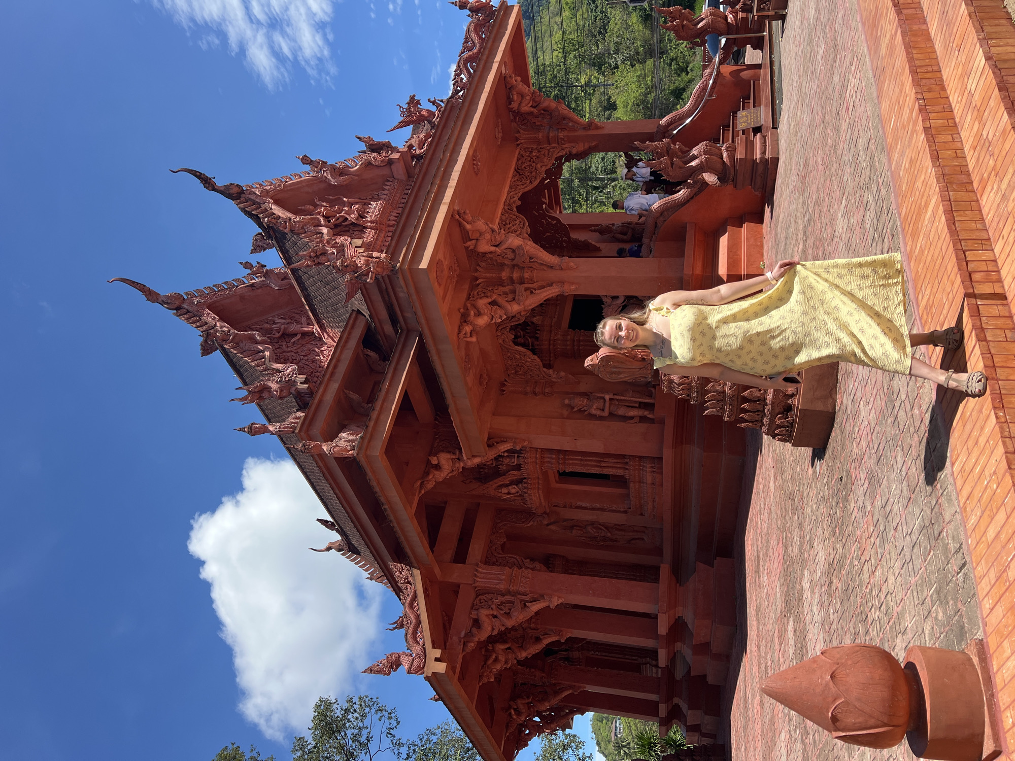 A red, ornate temple stands under a blue sky with scattered clouds. A woman in a yellow patterned dress stands on a brick tiled surface near the temple. The temple has intricate carvings and multiple tiers. Trees are visible in the background, suggesting a natural setting. The temple is located in Baan Hua Thanon, Thailand.