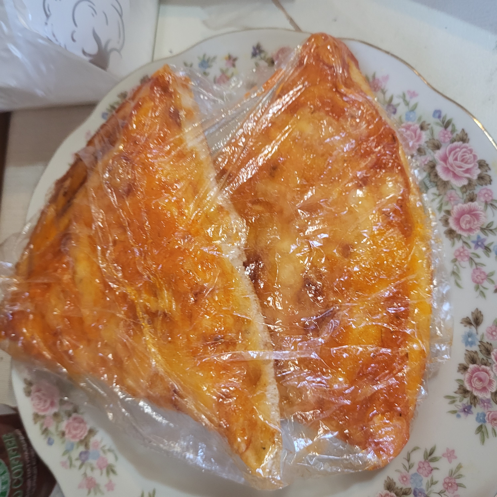 The image captures two pieces of golden-brown baked goods, likely a type of pastry or cheese bread, individually wrapped in clear plastic film. These items are placed on a decorative white plate with a floral pattern, featuring pink roses, blue and purple flowers, and green leaves, accented by a gold rim. The plate rests on a light-colored surface, suggesting an indoor setting such as a kitchen or dining area in Yerevan, Armenia.
The baked goods appear to be prepared for storage, transport, or later consumption, indicated by the plastic wrapping. In the upper left background, part of a white plastic bag or container with a faint gray design is visible. In the bottom left corner, a dark brown packet is partially obscured, but the word "COFFEE" is clearly legible on it. No people are visible, and there are no discernible cues for the time of day or weather.