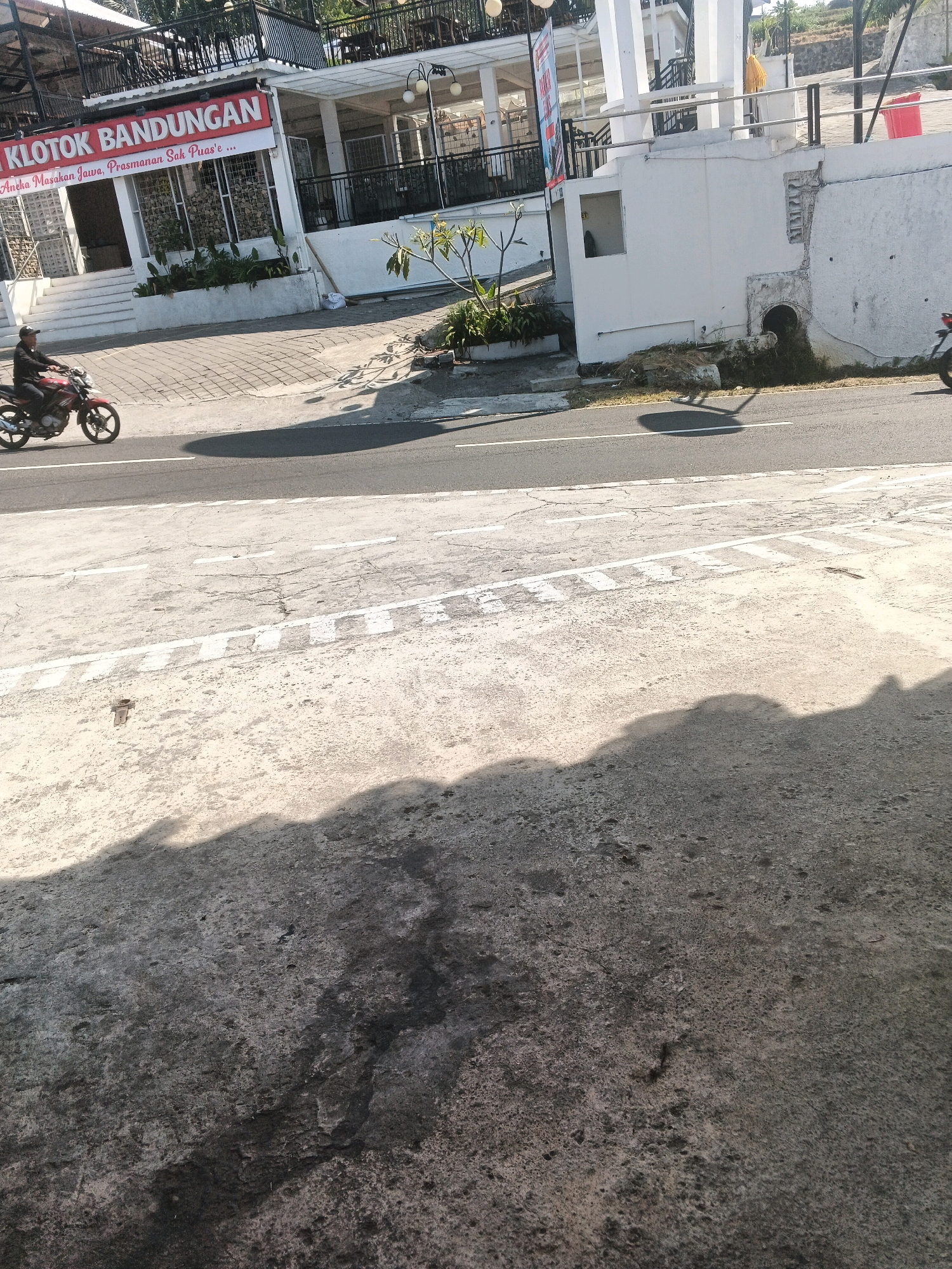 The image shows a scene in Pakopen, Indonesia, focusing on a road with a building and a motorcycle rider. On the left, a building with a red banner that reads "KLOTOK BANDUNGAN" is visible. Below the banner, text in Indonesian states "Aneka Masakan Jawa, Prasmanan Sak Puas 'e". A person is riding a red motorcycle on the road in front of the building. The road has white lines and a concrete shoulder with some vegetation. In the background, a white building with pillars and a railing can be seen, along with trees and a hill. The foreground of the image consists of the road and a concrete area with visible shadows, indicating a sunny day.