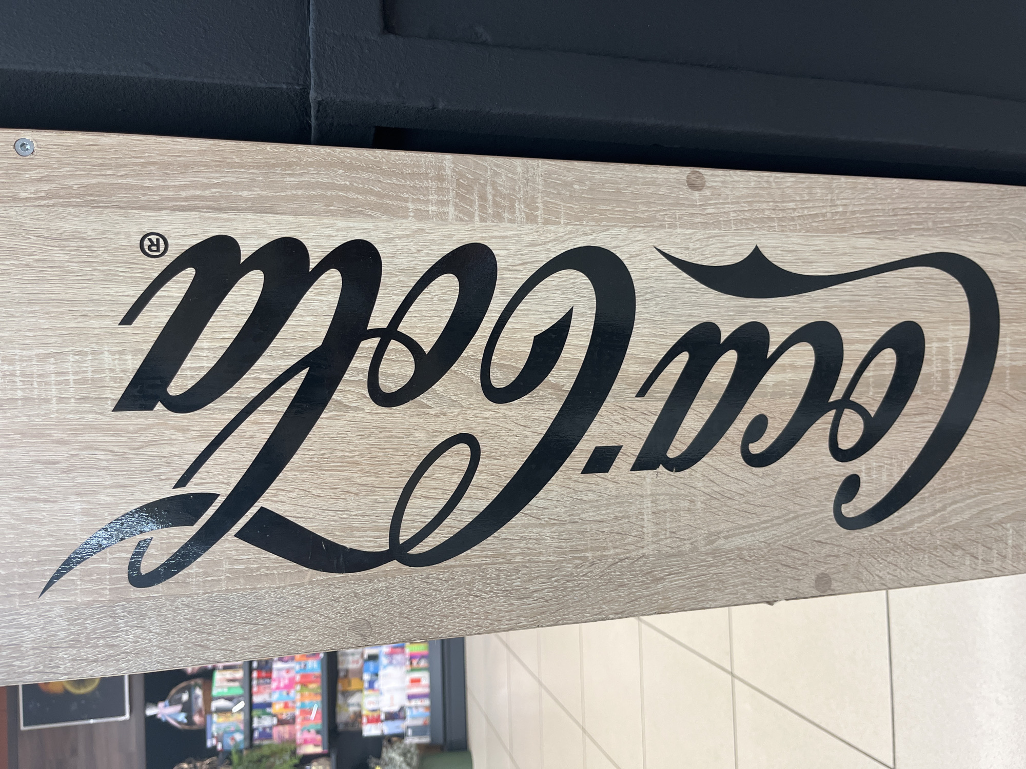 A vertically oriented, close-up shot displays the iconic Coca-Cola logo emblazoned in black script on a light-colored wooden surface. The logo begins at the top and extends downwards, with the stylized letters curving and flowing across the wood grain. A registered trademark symbol "®" is subtly integrated within the "C" of "Coca". To the right of the logo, a dark, textured surface, possibly a wall or another piece of furniture, forms a stark contrast. The background to the left reveals a blurred retail environment, with shelves stocked with colorful products and a tiled floor. The scene is likely from a shop or store in Kharkiv, Ukraine. There are no people visible in the image.