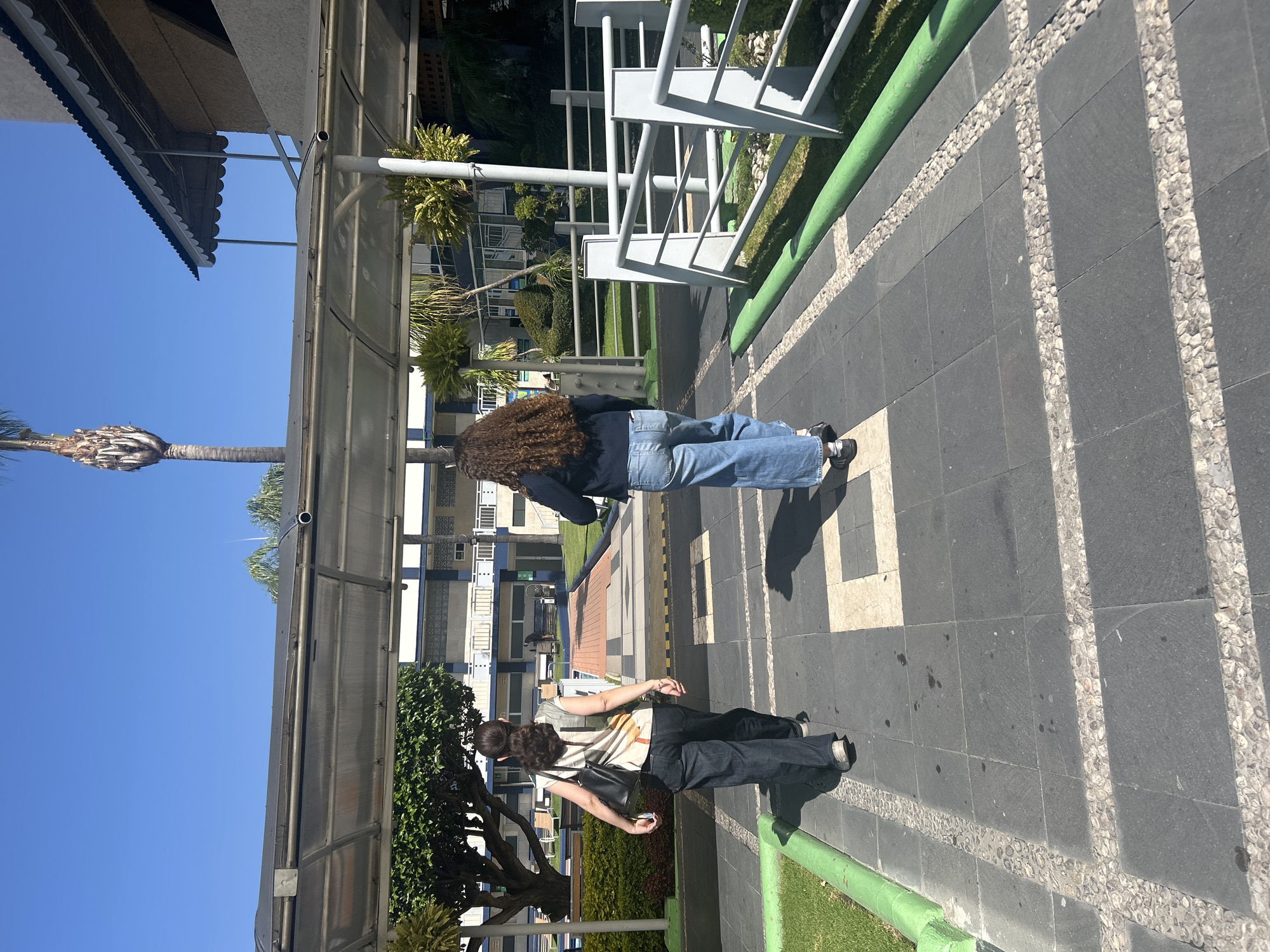 The image shows two individuals walking on a paved pathway outdoors. The person closer to the camera is wearing a striped shirt, dark pants, and carries a black bag. The other person is wearing a dark blue jacket and blue jeans. The pathway is made of dark grey tiles with lighter stone accents and runs alongside a green grassy area. A covered walkway structure stretches overhead, offering shade. Beyond the walkway, there are buildings and greenery, including palm trees. The sky is clear and blue.