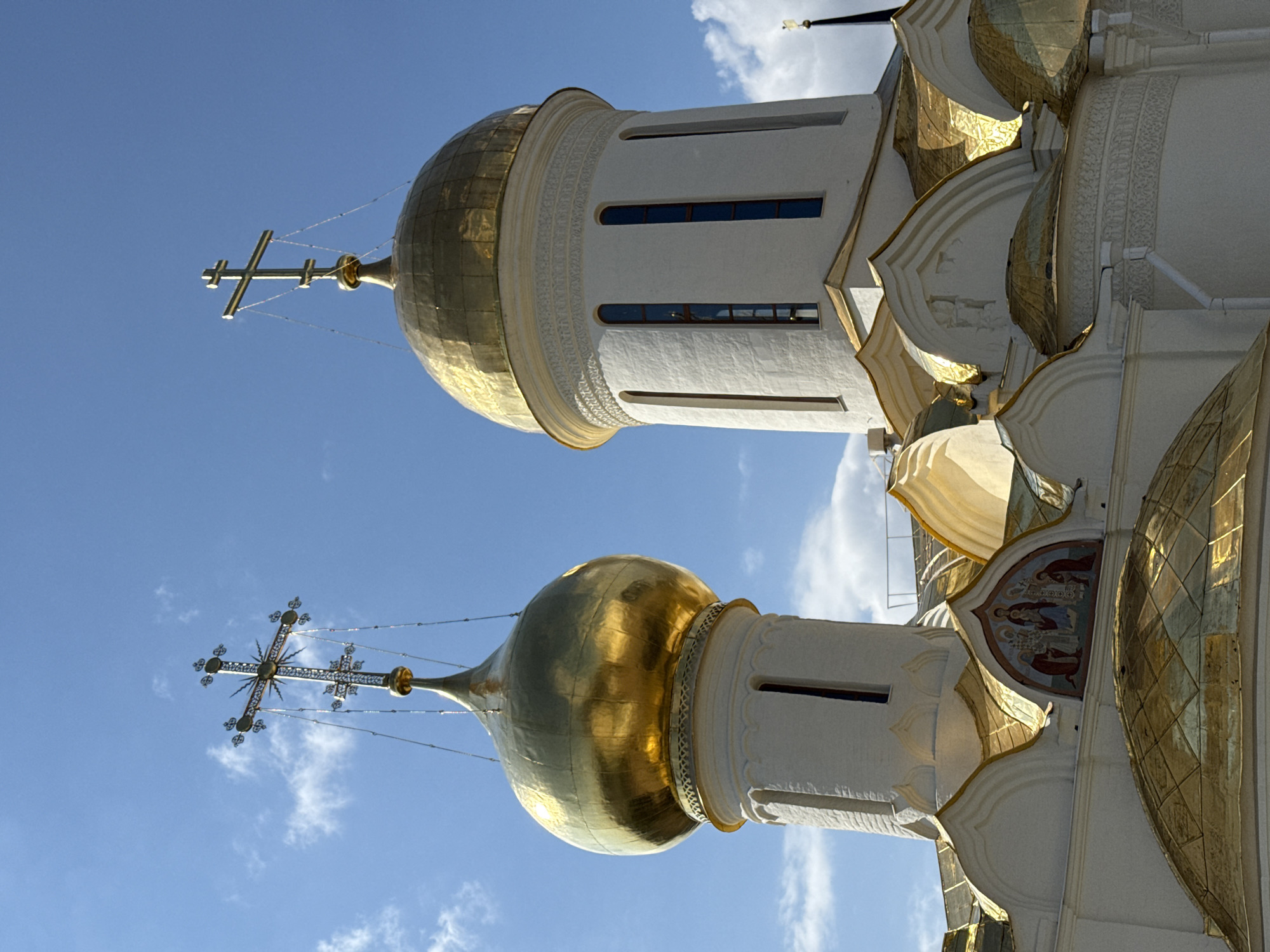 The image captures two golden domes of a church against a blue sky with scattered clouds. The domes have crosses on top. The church walls are white with gold trim. One section shows a religious mural. The location is Unknown, Unknown.