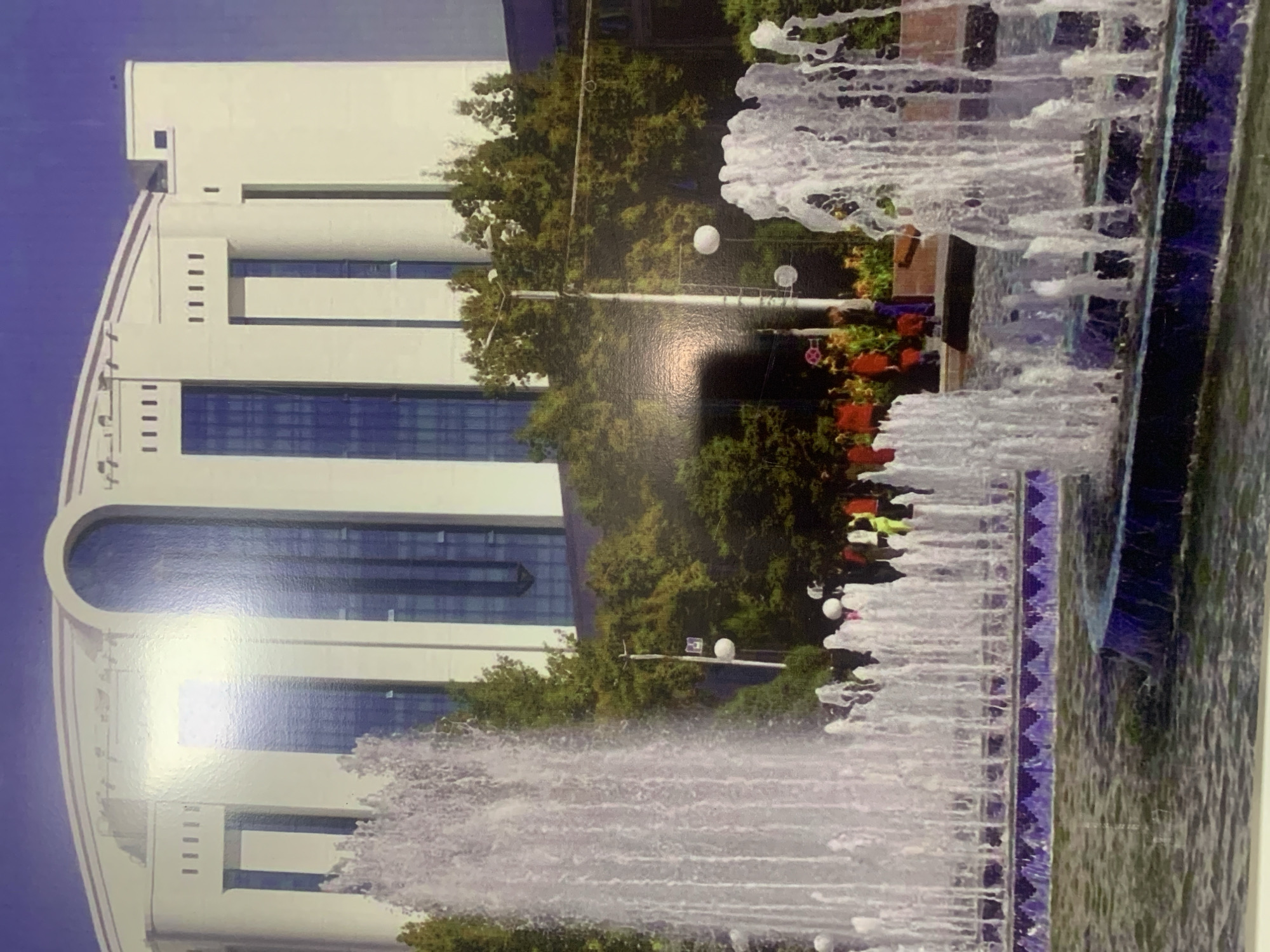 Here is a detailed description of the image:
The image depicts a large, white building with multiple floors and large windows. The architecture is modern with distinct lines and a curved top. In front of the building, there is a fountain with water spraying upwards. Trees and greenery are visible between the building and the fountain. There are people in the image, situated near the fountain. Streetlights are interspersed among the trees. The image is taken during daylight under a blue sky. The city location is Unknown, Unknown.