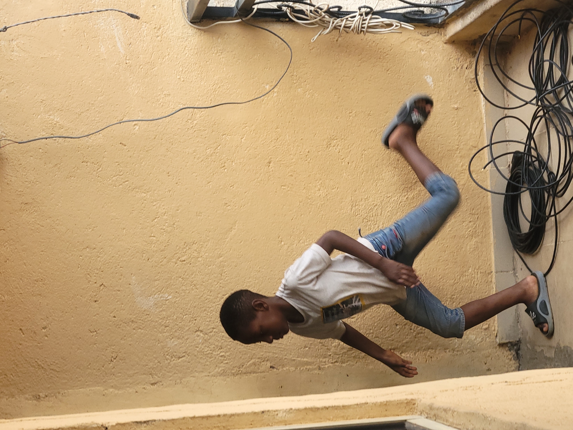 A photograph taken at an unknown location shows a young person in mid-air, seemingly falling or jumping, against a tan-colored wall. The individual, dressed in a white t-shirt and blue jeans, appears to be caught mid-fall, with one foot adorned in a gray sandal pointing towards the right side of the frame and the other raised higher. The surroundings include electrical wiring and cables positioned at the top of the wall, along with a section of a building corner visible on the right side. The texture of the wall is rough, suggesting a somewhat aged structure. A low wall segment is visible in the foreground.